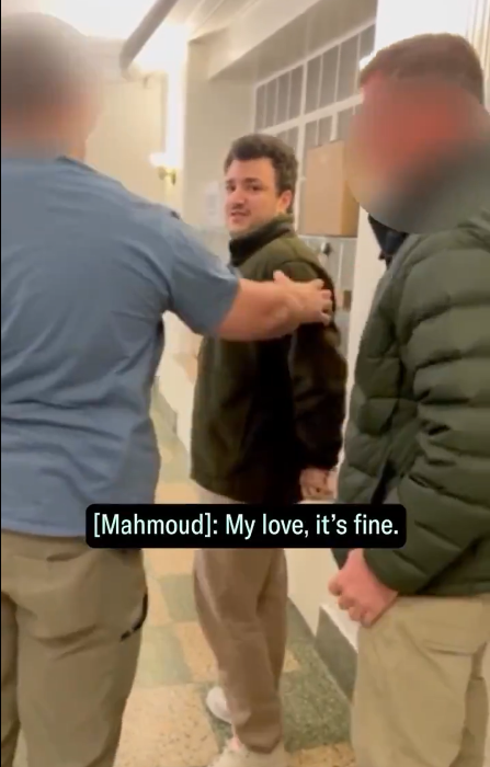 L'image montre un homme au centre, apparemment en train d'interagir avec deux autres hommes, dans un couloir. Un des hommes tient l'épaule de l'autre, et l'homme au centre semble parler, avec une expression préoccupée.