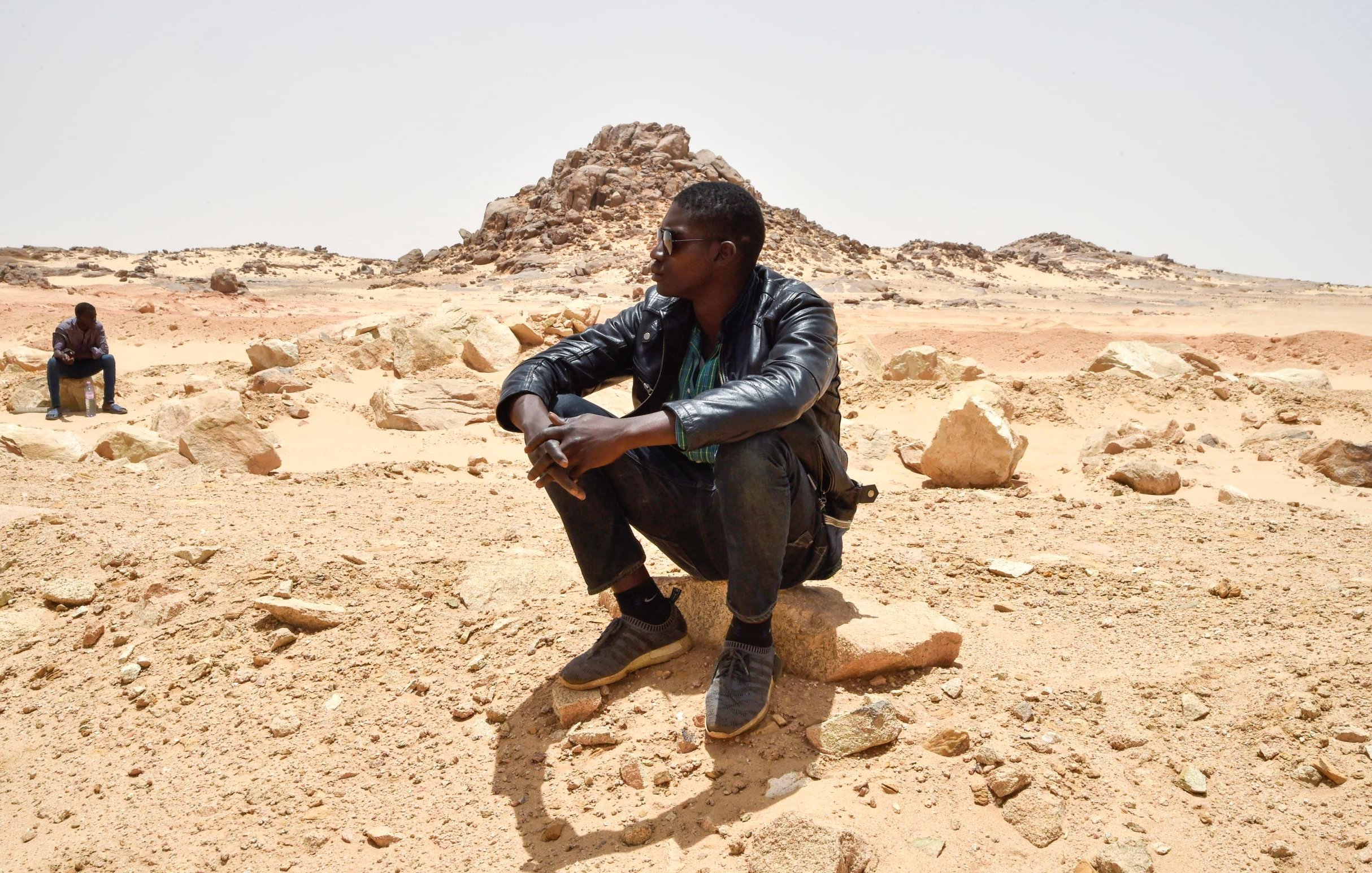 تظهر الصورة رجلين يجلسان في منطقة صحراوية. أحدهما يجلس على صخرة ويبدو عليه التفكير، بينما الآخر يجلس في الخلف بعيدًا قليلاً. الخلفية تتكون من صخور وكثبان رملية تحت سماء صافية. يبدو المشهد هادئًا، مما يعكس طبيعة الصحراء القاحلة.