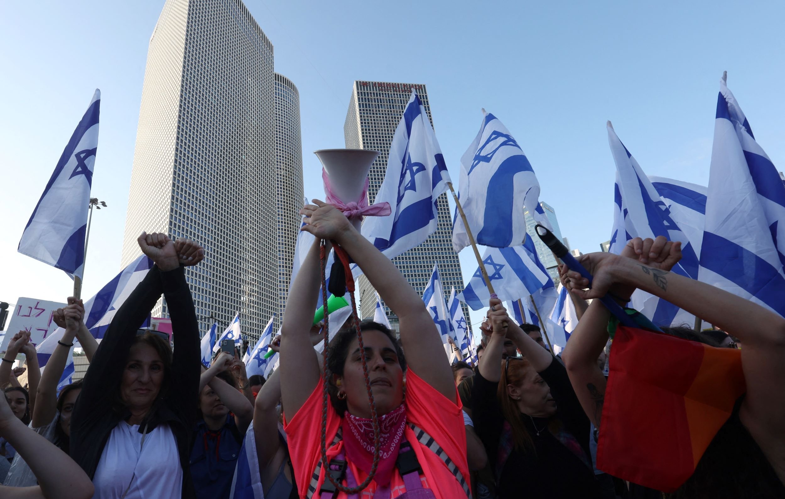 تظهر الصورة مجموعة من الأشخاص في مظاهرة، يحملون أعلامًا تحمل نجمة داود، وهو رمز معروف في إسرائيل. تظهر امرأة في المركز، مرفوعة اليدين وتحمل جهاز مكبر صوت، بينما يرفع الآخرون الأعلام. تتواجد خلفهم بنايات حديثة، مما يشير إلى أن المظاهرة قد تكون في منطقة حضرية. يعبّر المتظاهرون عن آرائهم، مما يدل على أهمية الفعالية التي يشاركون فيها.