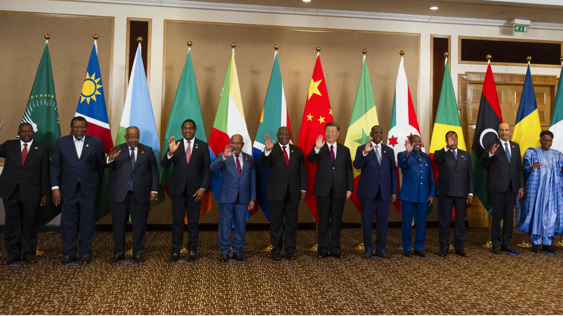 تظهر الصورة مجموعة من القادة أو الممثلين الرسميين من دول مختلفة يقفون في صف واحد. خلفهم، تُعلق أعلام تمثل الدول أو المنظمات المختلفة، بما في ذلك علم الاتحاد الأفريقي. يبدو أن الجميع يلوح بيديه في تعبير عن التحية. يرتدي القادة بدلات رسمية، وبعضهم يرتدي الملابس التقليدية. تعكس الصورة مناسبة رسمية أو قمة تجمع بين الدول.