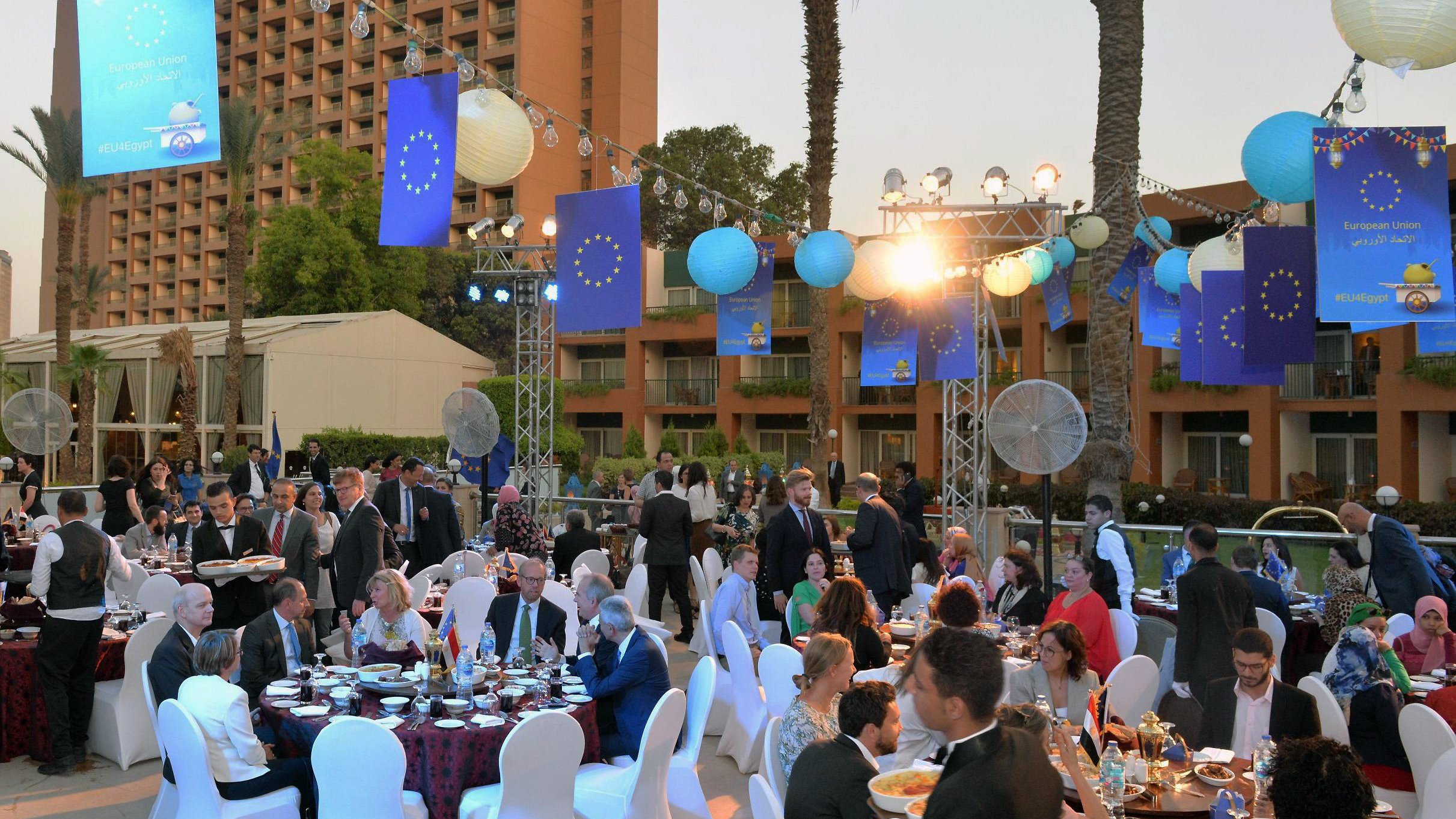 الصورة تظهر احتفالًا أو مناسبة اجتماعية في الهواء الطلق. هناك طاولات مزينة بأقمشة ملونة، وحولها مجموعة من الناس يرتدون ملابس أنيقة. في الخلفية، يبرز مبنى مرتفع، ويحيط بالمنطقة أشجار النخيل. الأضواء الداخلية والخارجية تعزز الأجواء الاحتفالية، وتوجد زينة على شكل كرات ملونة، بالإضافة إلى لافتات تحمل شعار الاتحاد الأوروبي. الأجواء تبدو مفعمة بالحياة والهدوء في الوقت نفسه.