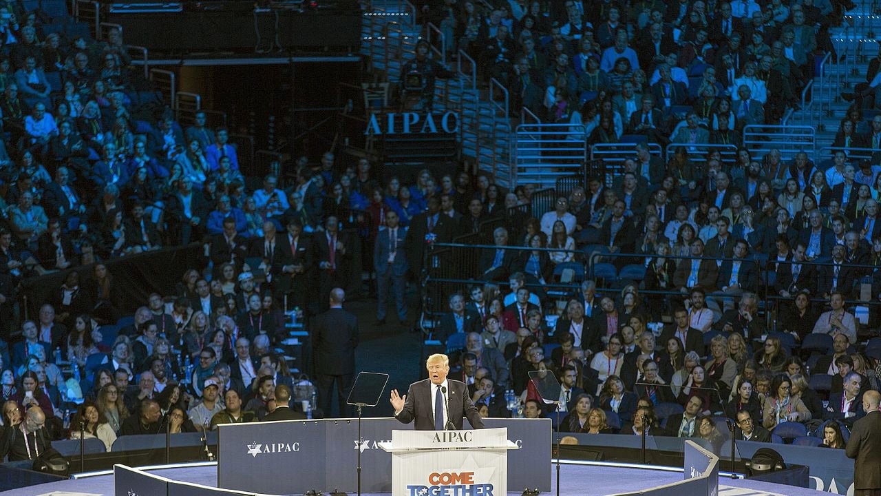 تظهر الصورة مشهداً لمؤتمر معبد مخصص لجمعية "AIPAC"، حيث يتحدث متحدث على المنصة أمام جمهور كبير. المنصة محاطة بأعلام وشعارات الجمعية، كما أن الإضاءة تلقي ظلالاً زرقاء على الحضور. يبدو أن المتحدث يناقش قضية مهمة، حيث يبدو الحضور متفاعلاً مع حديثه.