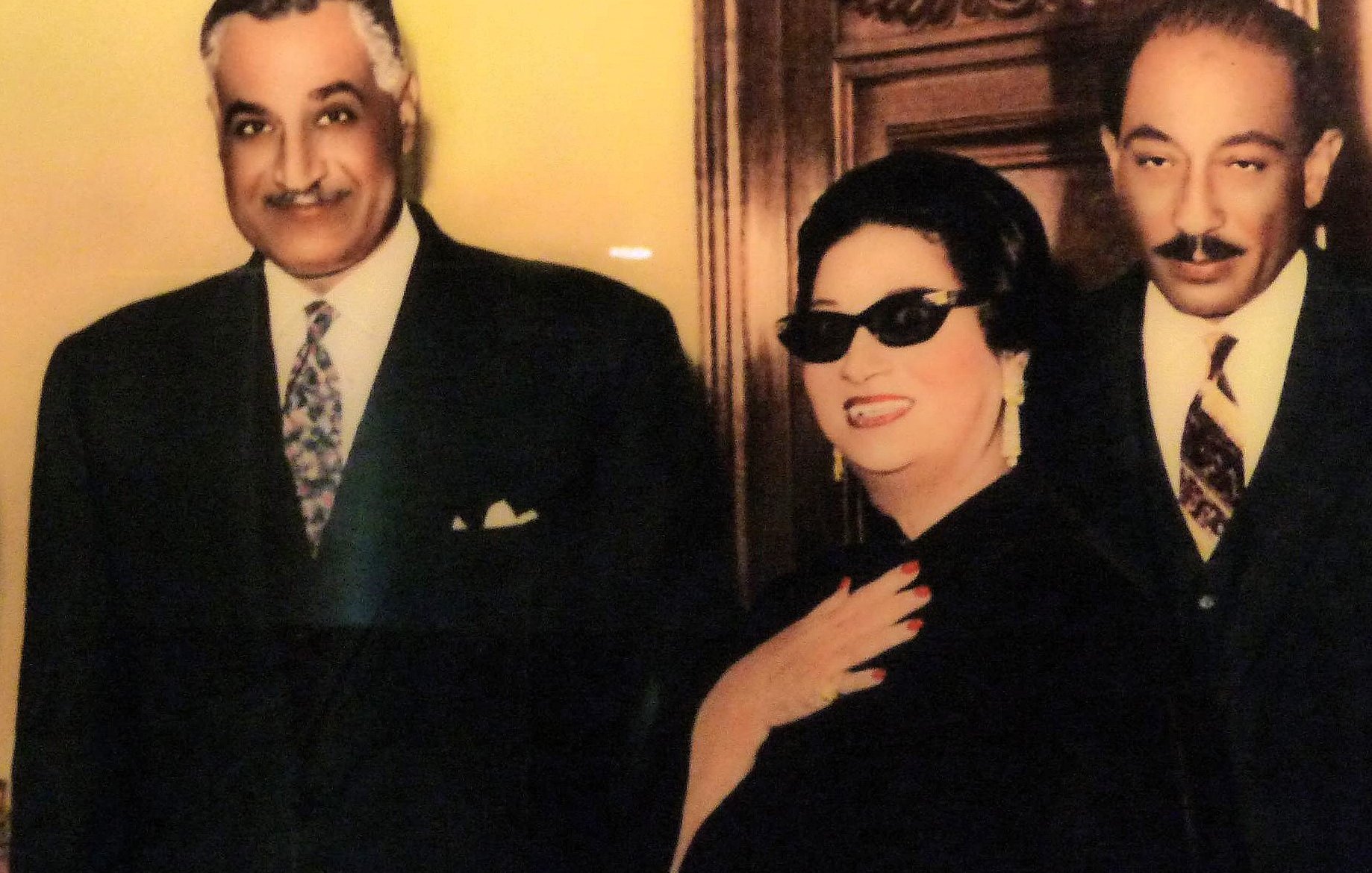 الصورة تظهر ثلاثة أشخاص يقفون معًا في إطار أنيق. الشخص الموجود على اليسار يبدو في بدلة رسمية وبابتسامة ودودة، بينما الشخصية في المنتصف ترتدي نظارات شمسية وقبعة، تبدو مميزة جدا في ملابسها، وتعبر عن شخصية قوية. الشخص الثالث على اليمين يبدو أيضًا في بدلة رسمية. الخلفية تحتوي على تصميم خشبي، مما يضيف لمسة من الفخامة إلى الصورة.