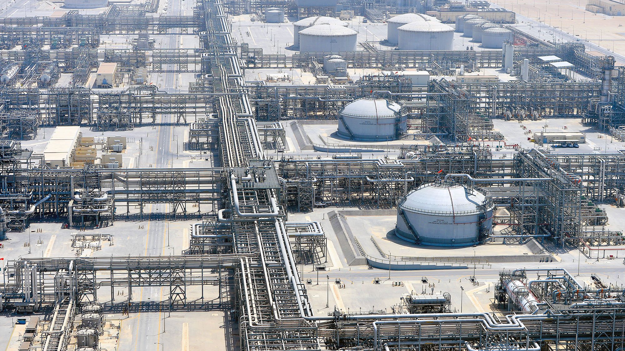 الصورة تُظهر منظرًا جويًا لمصنع صناعي ضخم، يبدو أنه يتعلق بقطاع النفط أو الغاز. يمكن رؤية مجموعة من الخزانات الدائرية الكبيرة، بالإضافة إلى شبكة معقدة من الأنابيب والبنى التحتية الأخرى. المنطقة محاطة بأرض صحراوية، مما يدل على أن المصنع يقع في منطقة جافة. يركز التصميم على الكفاءة في نقل وإدارة المواد الخام، مما يشير إلى مستوى عالٍ من التكنولوجيا والهندسة.