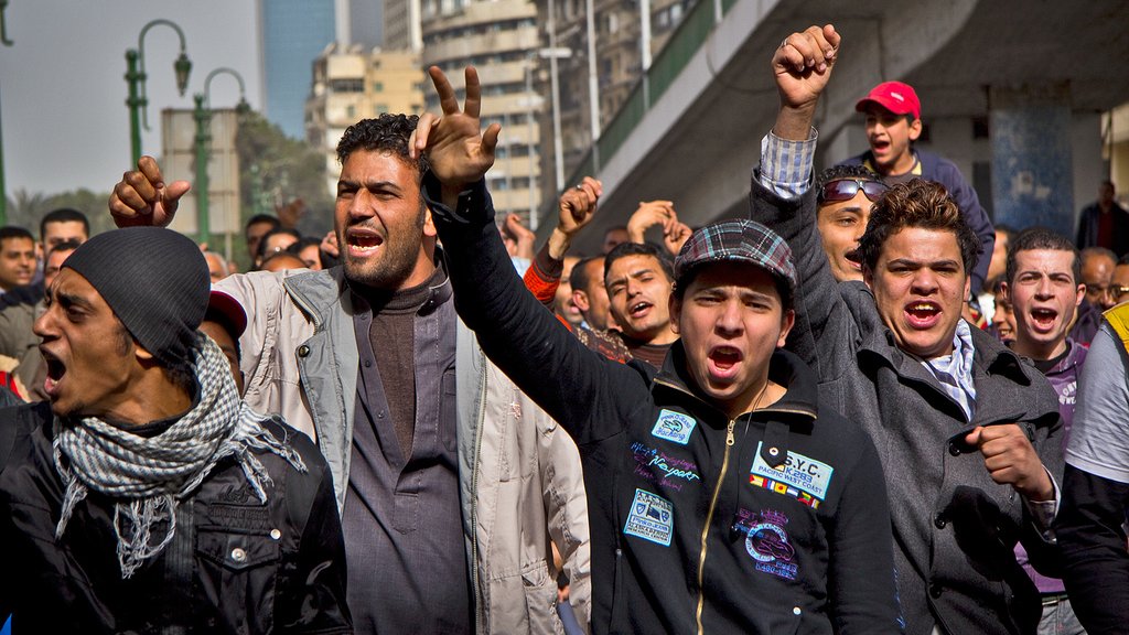 تظهر في الصورة مجموعة من الشباب الذين يقفون في منطقة حضرية. يبدو أنهم يعبرون عن مشاعر قوية، حيث يرفعون قبضاتهم وينظرون إلى الأمام بحماس. تعكس تعابير وجوههم الحماسة أو الغضب، وقد يكونون في حالة احتجاج أو تجمع جماهيري. يحيط بهم مبانٍ عالية وعناصر حضرية، مما يدل على أن النشاط يحدث في مدينة.