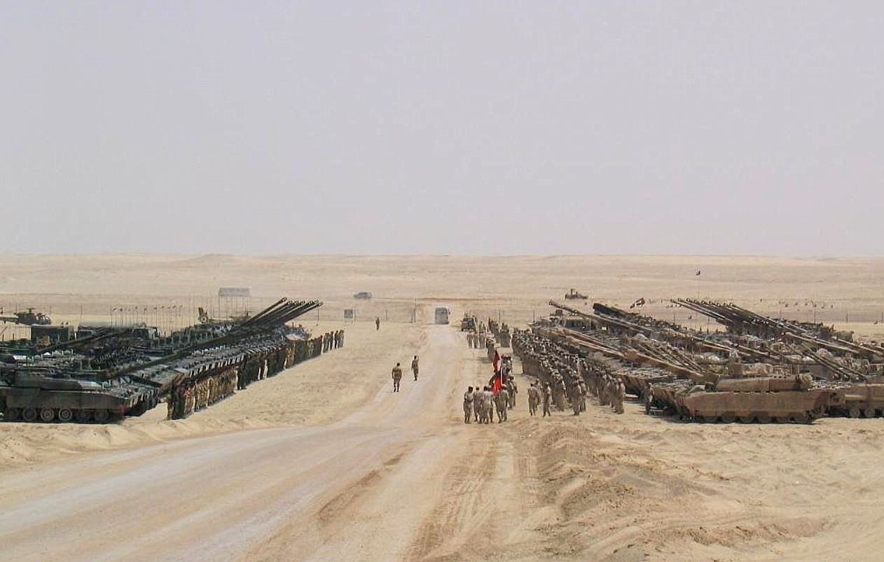 تظهر في الصورة مشهدًا صحراويًا يضم العديد من الدبابات والمركبات العسكرية. يقف عدد من الجنود في المنتصف، بينما تتواجد مركبات أخرى مصفوفة على جانبي الطريق. الخلفية تتكون من رمال الصحراء وأفق بعيد، مما يبرز الطبيعة القاحلة للمكان. تخلق الصورة انطباعًا بالقوة العسكرية والتنظيم.