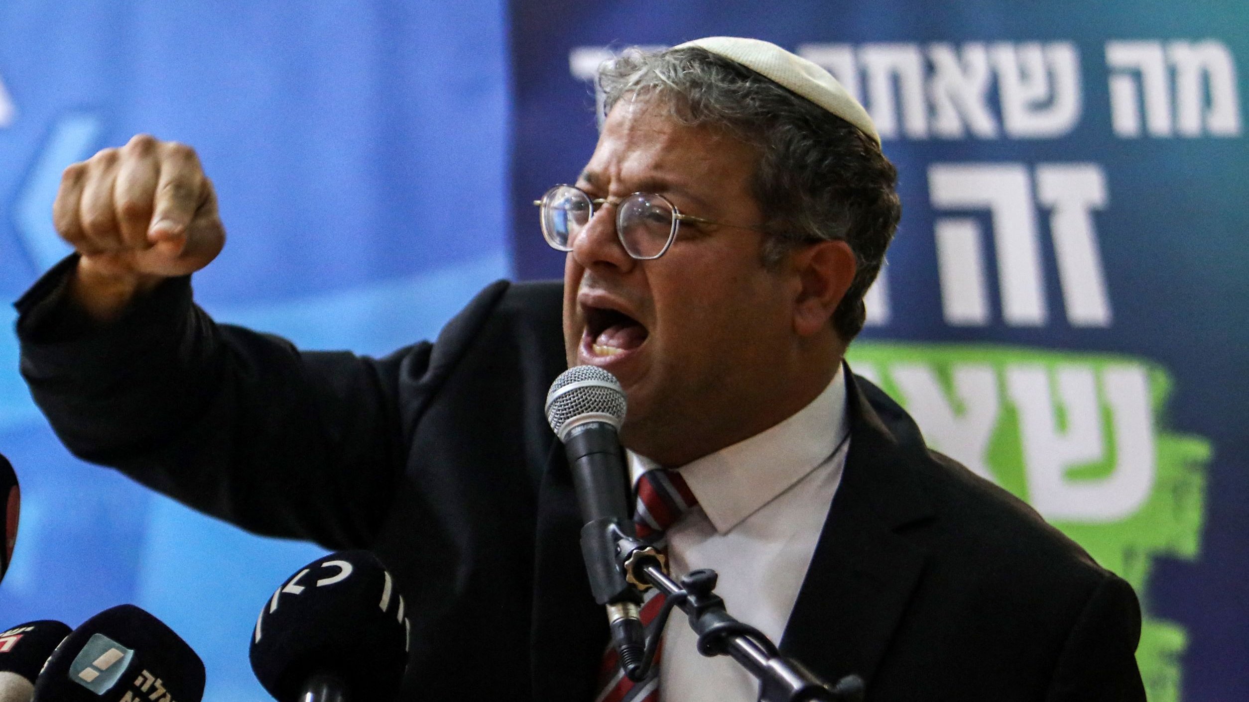 تظهر الصورة شخصية تتحدث بحماسة أمام ميكروفون خلال مؤتمر أو حدث عام. يرتدي بدلة رسمية ونظارات، ويبدو أنه يعبر عن مشاعر قوية، حيث يشير بيده ويظهر تعابير وجه تعكس الحماس أو الغضب. الخلفية تحتوي على شعارات أو نصوص، مما يدل على أن الحدث قد يكون سياسيًا أو يتعلق بقضية عامة.