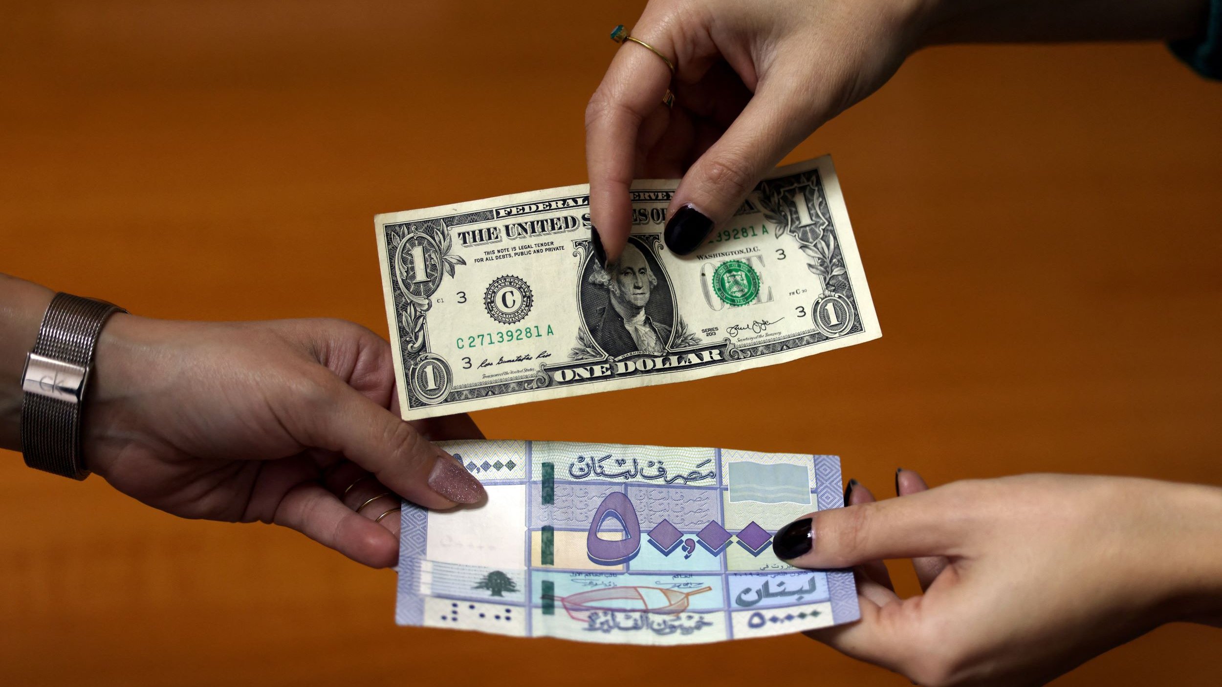 تظهر في الصورة يدان تتبادلان النقود، حيث تحمل يد واحدة دولارًا أمريكيًا من فئة الواحد دولار، بينما تحمل اليد الأخرى عملة محلية. تتوسط الأيدي خلفية خشبية، مما يعكس عملية تبادل أو صفقة.