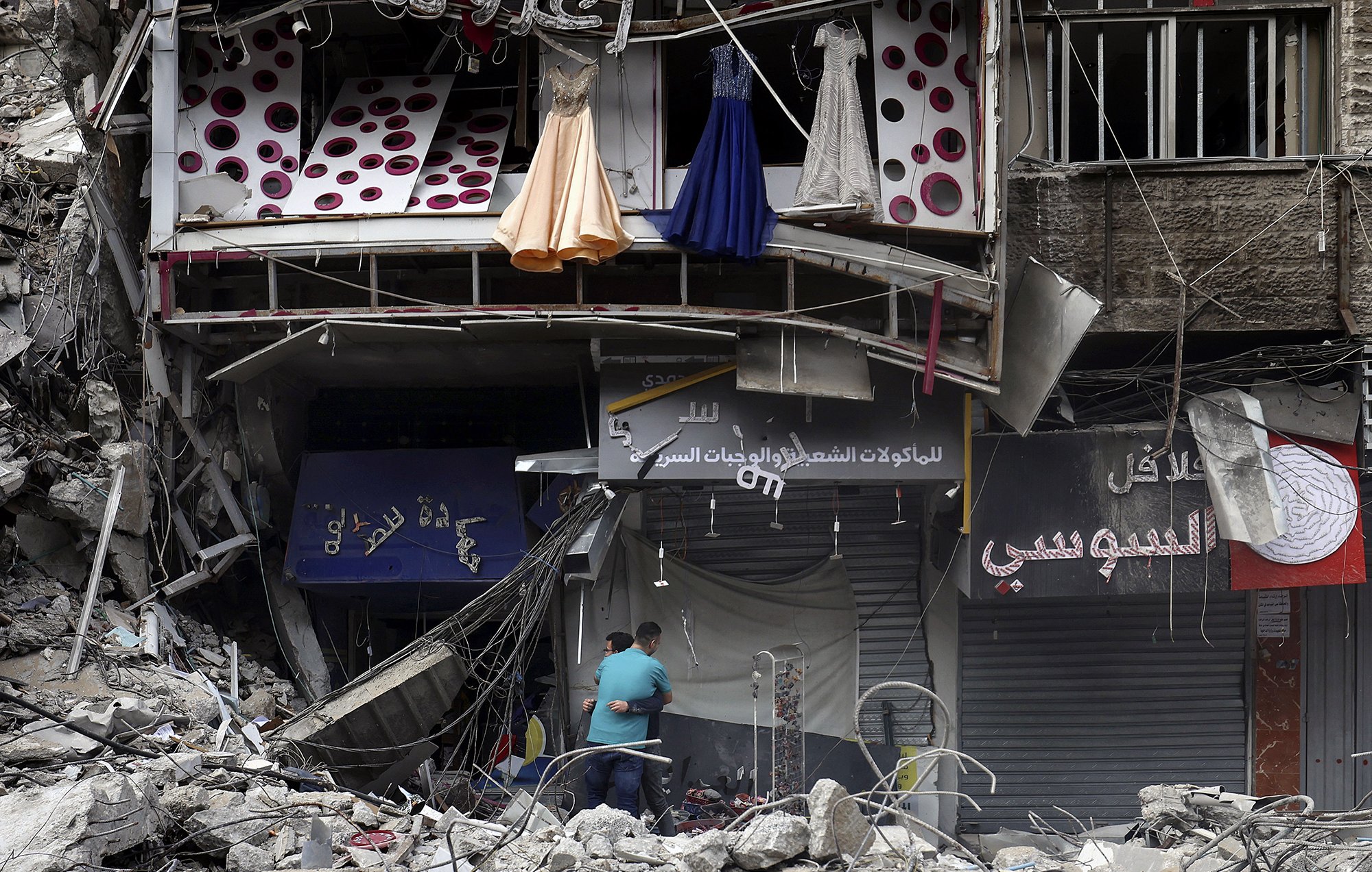 تظهر الصورة مشهدًا من منطقة دمار. هناك مبنى مهدوم جزئيًا يظهر فيه بعض الملابس المعلقة، مثل فساتين، بالإضافة إلى واجهات متاجر متضررة. يلاحظ في الأسفل شخصان يتفقدان المكان وسط الأنقاض. المشهد يعكس آثار تدمير كبير والخراب الذي لحق بالمنطقة.