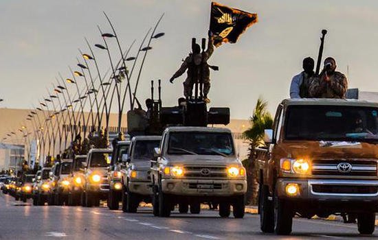 تظهر الصورة قافلة من الشاحنات تسير في شارع، حيث يوجد عدد من الأشخاص المسلحين على متنها. يُظهر جزء من القافلة لافتة سوداء تُشير إلى رمزية معينة. تبدو الأجواء في الصورة مائلة إلى المساء، مع وجود أعمدة إنارة على جوانب الطريق. توحي الصورة بحركة عسكرية أو تحركات لمجموعة معينة.