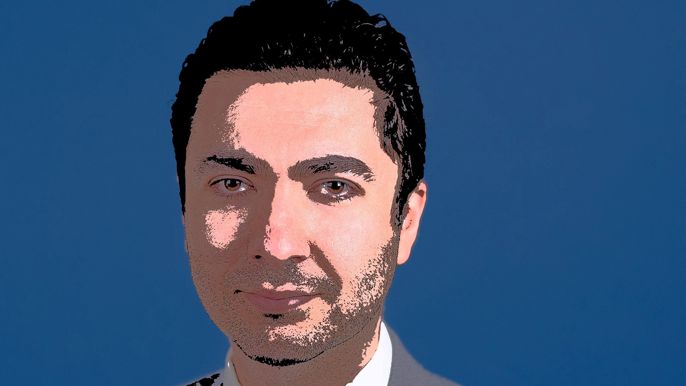 الصورة تظهر شخصاً يرتدي بدلة رسمية باللون الرمادي، مع قميص أبيض وربطة عنق زرقاء. الخلفية ذات لون أزرق، ويبدو على وجهه تعبير واثق. الشعر داكن وممشط بشكل مرتب.