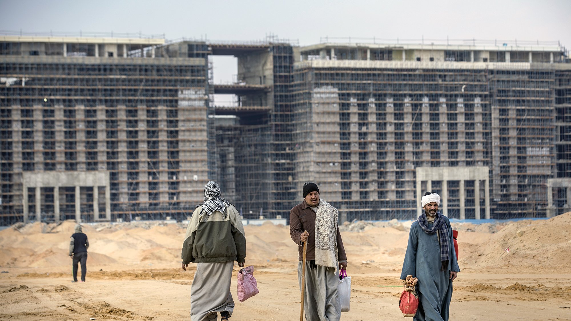 في الصورة نرى مجموعة من الأشخاص يمشون على طريق رملي في منطقة قيد الإنشاء. خلفهم توجد مبانٍ مرتفعة لا تزال في مراحل البناء، مع وجود هياكل معدنية وأخشاب واضحة. يرتدي الأشخاص ملابس تقليدية، ويبدو البعض منهم يحمل حقائب. السماء ملبدة بالغيوم، مما يعطي انطباعًا بجو من الصبر والعمل الجاد في بيئة الإنشاءات.