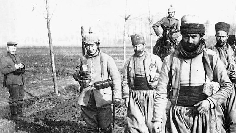 الصورة تظهر مجموعة من الجنود يسيرون في منطقة مفتوحة، يبدو أن الأرض مبللة ومليئة بالطين. الجنود يرتدون زيًا عسكريًا تقليديًا، بعضهم يحمل أسلحة، بينما يبدو أحدهم راكبًا على حصان. يمكن رؤية شخص آخر في الخلفية يقف على الجانب. الأجواء العامة توحي بأنها قد تكون خلال فترة تاريخية معينة من الحرب أو النزاع.