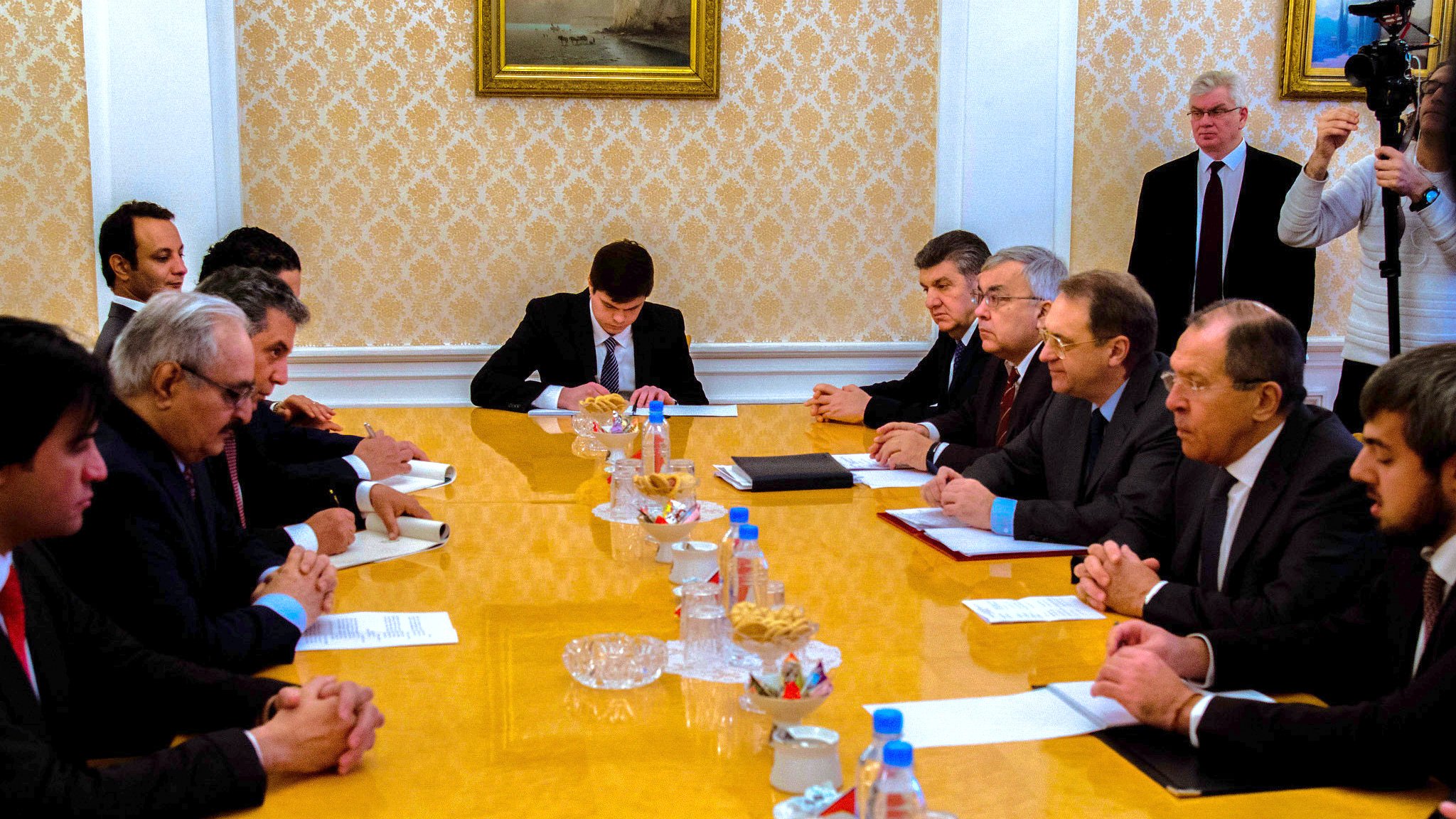 تظهر الصورة اجتماعاً رسمياً يتم فيه نقاش أو تبادل للأفكار بين مجموعة من الأشخاص. يجلس المشاركون حول طاولة كبيرة تحتوي على مستندات وبعض الوجبات الخفيفة. يبدو أن هناك تركيزاً على النقاش، حيث يظهر البعض وهم يكتبون أو يتطلعون إلى المستندات الموجودة أمامهم. الخلفية تحتوي على لوحات فنية تزين الجدران، مما يضيف طابعاً رسمياً إلى الاجتماع.