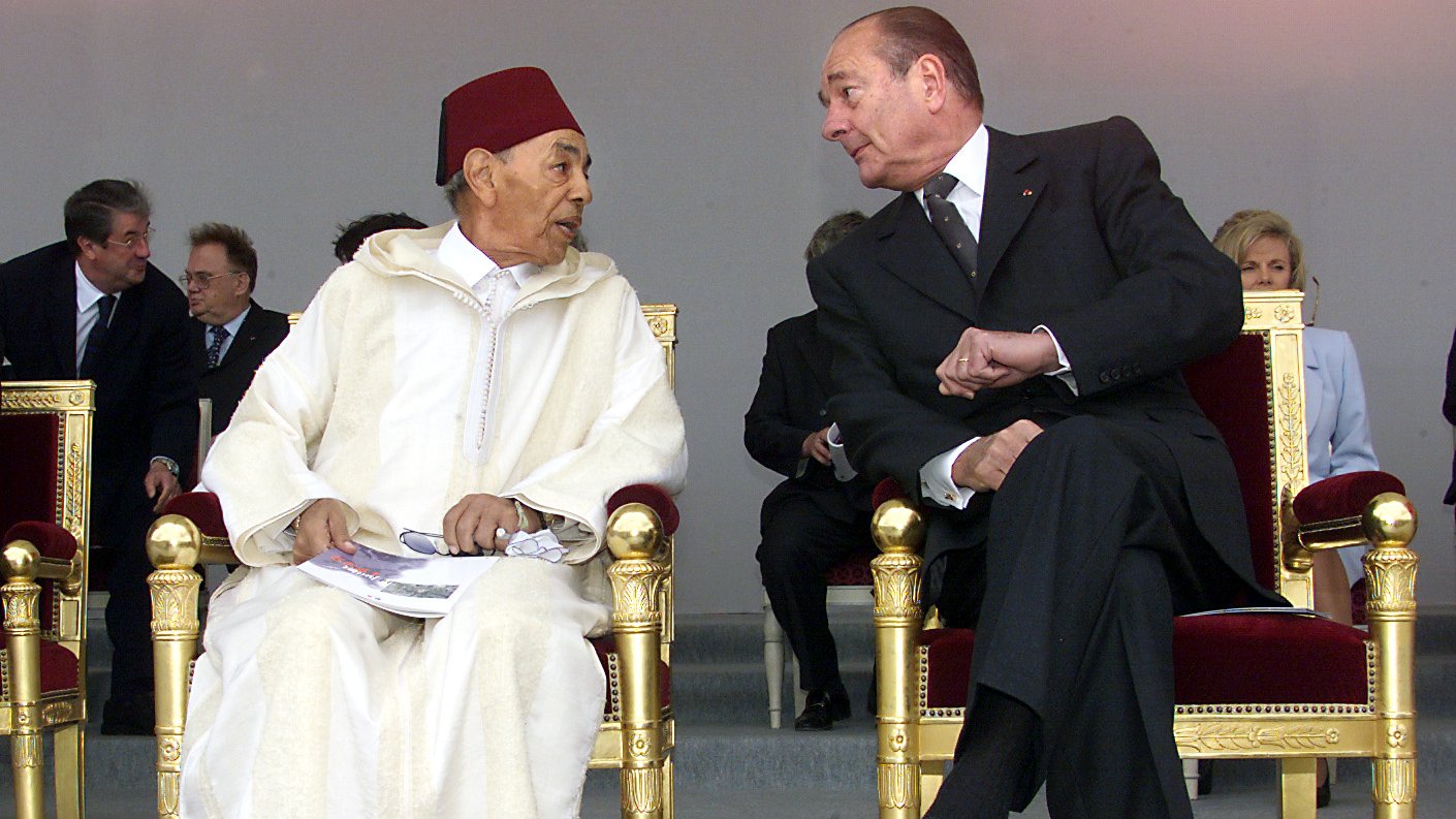 الصورة تظهر شخصين يجلسان على كراسي فاخرة. يبدو أن الحديث يجري بينهما بشكل ودي. الشخص الأول يرتدي زيًا تقليديًا مغربيًا مع طربوش، بينما الشخص الثاني يرتدي بدلة رسمية داكنة. في الخلفية توجد مجموعة من الأشخاص الجالسين، ويبدو أنهم في مناسبة رسمية أو احتفال. الضوء في الصورة ناعم، مما يضيف جوًا من الألفة.