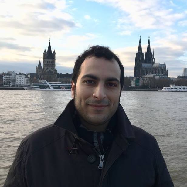 L'image montre un homme se tenant au bord d'un fleuve, probablement le Rhin, avec un paysage urbain en arrière-plan. On peut voir des bâtiments emblématiques, dont une grande cathédrale avec des flèches pointues et une architecture gothique. Le ciel est nuageux avec quelques éclaircies, ce qui donne une ambiance calme à la scène. L'homme porte un manteau sombre et affiche un léger sourire.