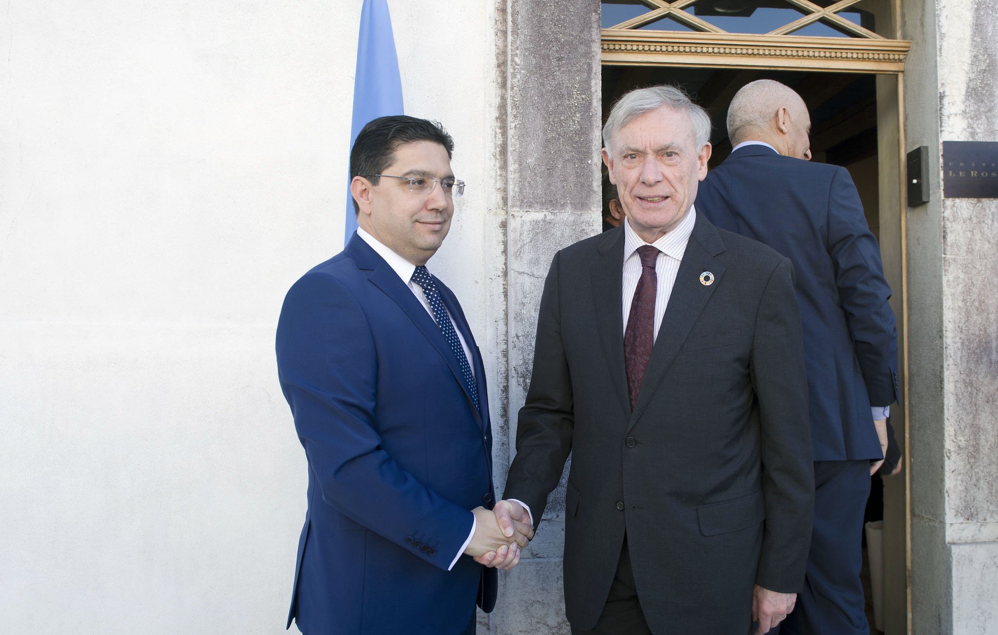 الصورة تظهر شخصين يتصافحان خارج مبنى. الشخص الأول يرتدي بدلة زرقاء ويبدو عليه الجدية. الشخص الثاني يرتدي بدلة داكنة ويظهر تعبيراً ودياً. في الخلفية، يمكن رؤية بعض الأشخاص الآخرين يدخلون أو يغادرون المكان، بالإضافة إلى العلم الذي يظهر في الزاوية.