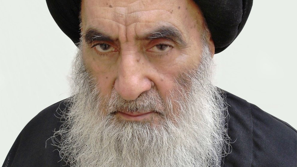 الصورة تُظهر رجلًا مسنًّا ذو لحية بيضاء وشعر داكن تحت عمامة سوداء. يبدو عليه الوقار والجدية. يرتدي ثوبًا داكنًا، مما يعكس نمطًا تقليديًا في اللباس. الخلفية بسيطة ووردية اللون، مما يبرز ملامح وجهه.