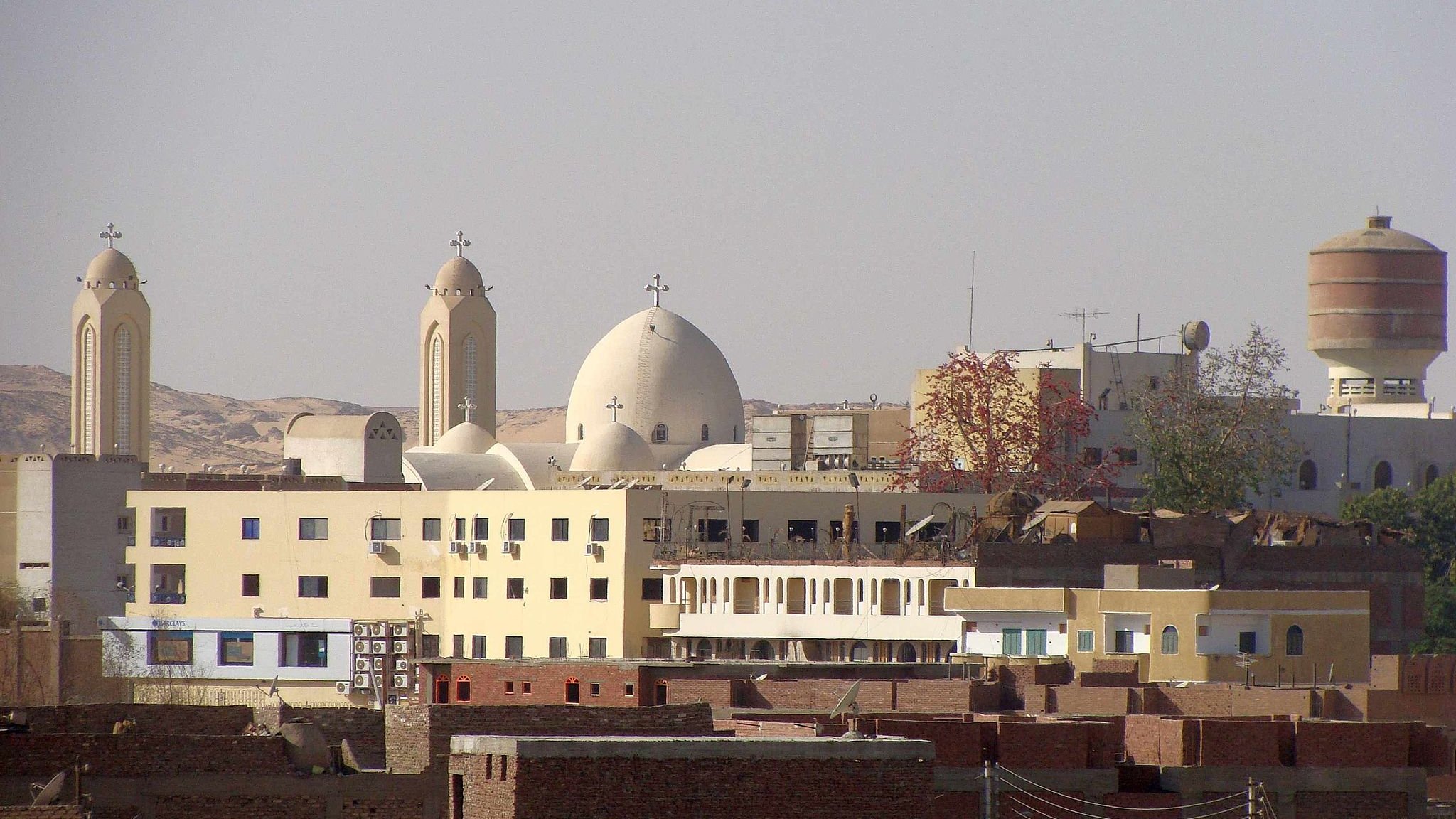 الصورة تظهر مجموعة من المباني في منطقة حضرية، حيث يتواجد مباني ذات طراز معماري مميز، بما في ذلك الكنائس التي تتضمن قبابًا وجرسًا. الخلفية تحتوي على تلال وصحراء، مما يوحي بأن المنطقة قد تكون قريبة من الطبيعة. يوجد أيضًا مجموعة من المباني السكنية المنخفضة، تظهر على اليمين، مما يعطي انطباعًا عن الحياة اليومية في تلك المنطقة.