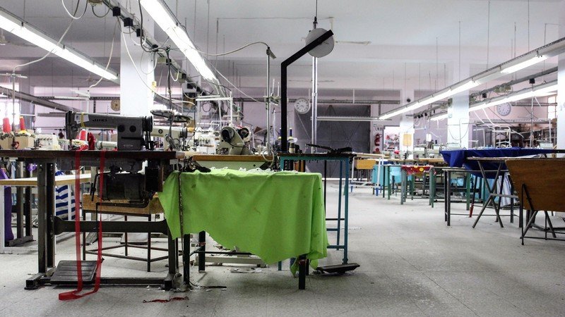 تظهر الصورة مشهدًا داخليًا لورشة خياطة. المساحة تبدو واسعة ومشرقة، حيث توجد آلات خياطة متعددة منتشرة في المكان. بعض الطاولات تحتوي على أقمشة ملونة، في حين أن العديد من المعدات والأدوات موجودة في حالة استخدام أو جاهزية. الأرضية مغطاة ببعض القصاصات من القماش، مما يشير إلى نشاط العمل. الإضاءة تأتي من الأنوار السقفية، وهناك ساعة على الحائط تدل على الوقت. يبدو أن الورشة تحتاج إلى بعض الترتيب.