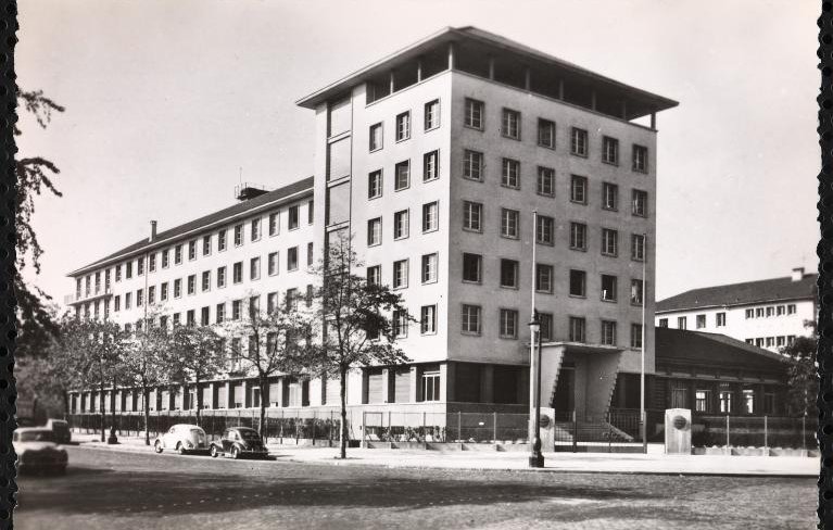 الصورة تُظهر مبنىً حديث التصميم، يتكون من عدة طوابق وله واجهة متنوعة الألوان. يتواجد حول المبنى مجموعة من الأشجار، ويبدو أن هناك شارعًا أمام المبنى حيث توجد بعض السيارات القديمة. يعكس تصميم المبنى طابعًا معماريًا بسيطًا وعصريًا، مما يوحي بأنه تم بنائه في فترة زمنية معينة في القرن العشرين. السماء تبدو واضحة مما يعطي انطباعًا بأن الصورة قد تم التقاطها في يوم مشمس.