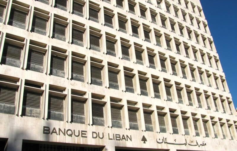 الصورة تظهر واجهة مبنى كبير يتكون من عدة طوابق، ويتميز بتصميم معماري حديث وبسيط. الجدران مصنوعة من الحجر أو الخرسانة، مع نوافذ موزعة بشكل منتظم. يظهر اسم البنك "BANQUE DU LIBAN" باللغة الفرنسية، مع إضافة عبارة باللغة العربية "مـصرف لبنان" تحتها. المبنى يعكس الطابع المؤسسي ويبدو أنه يقع تحت ضوء الشمس، مما يعطي شعوراً بالوضوح والنقاء.