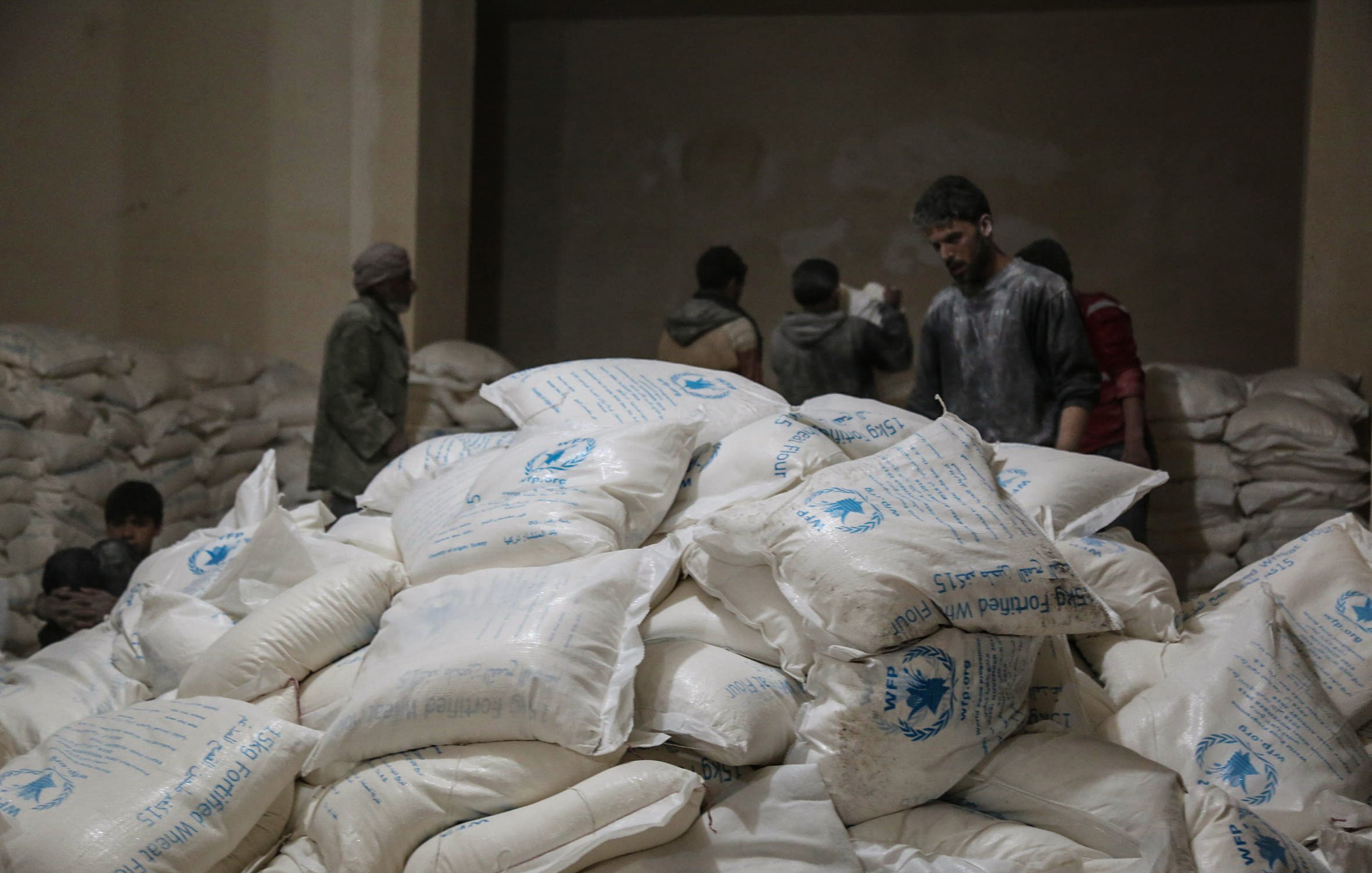 تظهر في الصورة مجموعة من الأشخاص داخل مكان يبدو أنه مخزن للمواد الإغاثية. في المقدمة، هناك كومة كبيرة من الأكياس البيضاء التي تحمل علامات معينة، ويبدو أن الأكياس تحتوي على مواد غذائية أو إغاثية. في الخلفية، يمكن رؤية بعض الأشخاص الذين يتحدثون أو يعملون. الإضاءة في المكان خافتة، مما يعطي شعورًا بالجدية والاهتمام بالعمل الذي يقومون به.