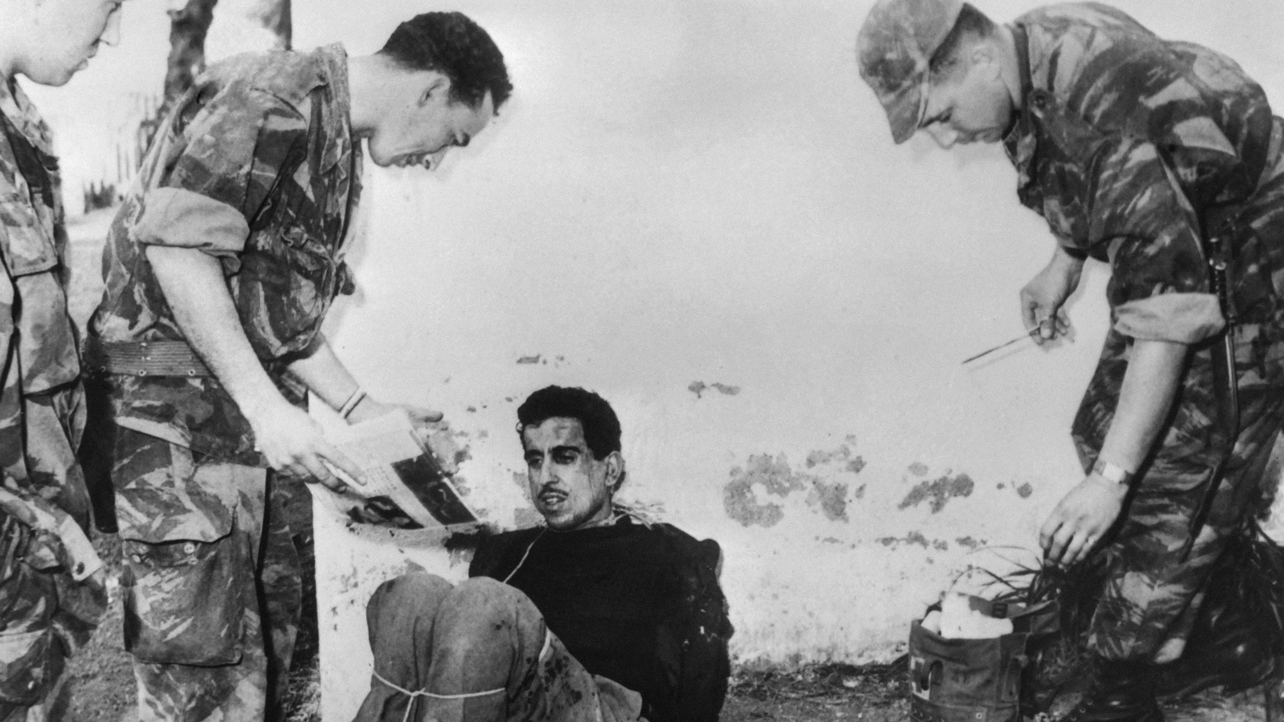 الصورة تظهر مجموعة من الجنود أثناء استجواب رجل يبدو أنه محتجز. الرجل جالس على الأرض ومربوط. الجنود يظهرون بزيهم العسكري، ويبدو أنهم يركزون على الوثائق أو الصور. البيئة المحيطة تبدو خارجية وملوثة بعض الشيء، مما يوحي بمكان غير رسمي. تعكس الصورة أجواء توتر وصعوبة.