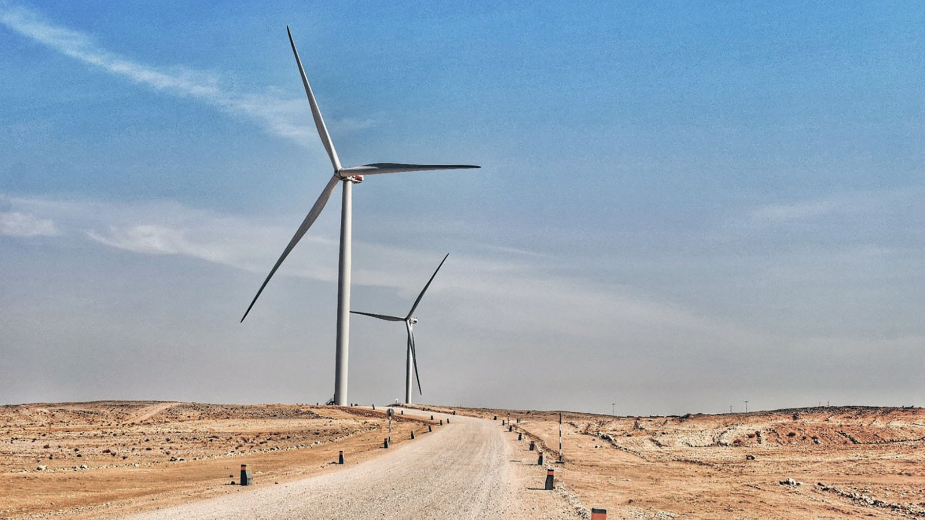 تظهر في هذه الصورة منظرًا طبيعيًا صحراويًا يمتد تحت سماء زرقاء فاتحة. يتواجد في الجزء الأمامي طريق رملي يتجه نحو الأفق، محاطًا بأعمدة صغيرة على جانبيه. في الخلفية، هناك عدة توربينات رياح ضخمة تدور ببطء، مما يُعطي انطباعًا حديثًا حول استخدام الطاقة المتجددة في هذا المناطق القاحلة. يبدو المكان هادئًا وواسعًا، مع تباين واضح بين الألوان الرمادية للأرض والألوان الزرقاء للسماء.