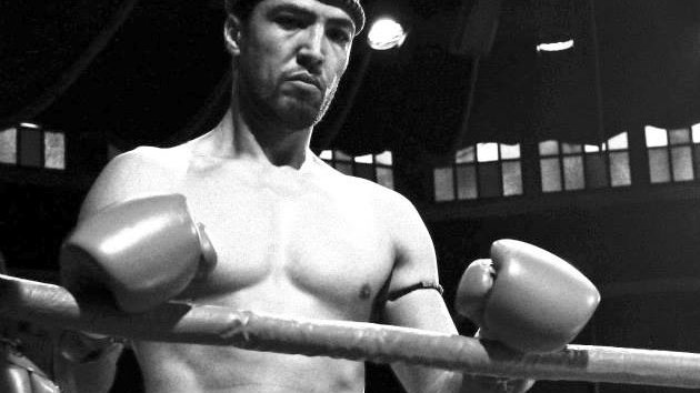 الصورة تظهر الملاكم في حلبة الملاكمة، حيث يبدو عابسًا وجادًا. هو يرتدي حزامًا أو شورت رياضي مكتوب عليه "RAJA" وتظهر على يديه قفازات الملاكمة. تعكس الإضاءة ونمط الصورة باللونين الأبيض والأسود أجواء المنافسة والقتال.