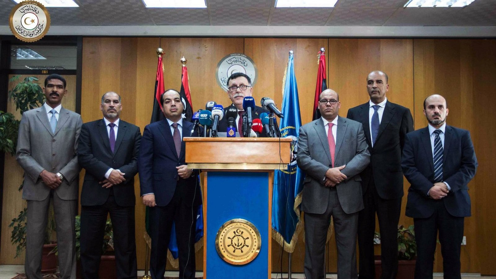 الصورة تظهر مجموعة من الرجال الواقفين في قاعة مؤتمرات. يتوسطهم شخص يقف أمام منصة تحمل شعار رسمي، وهو يبدو وكأنه يتحدث أو يقدم بيانًا. الخلفية تحتوي على أعلام متعددة، وقد تكون إشارة إلى مناسبة رسمية أو مؤتمر. جميع الحضور يرتدون بدلات رسمية ويظهر عليهم الجدية.