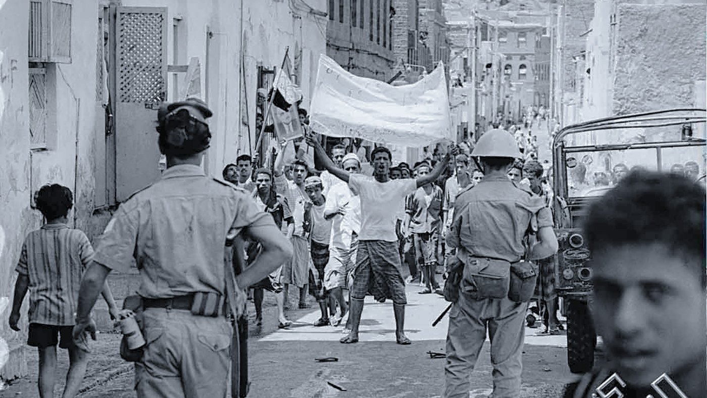 تظهر الصورة مجموعة من الأشخاص في شارع ضيق، يبدو أنهم في خضم حدث أو تظاهرة. في المقدمة، هناك رجل يرفع يديه حاملاً لافتة، بينما يتواجد أفراد من قوات الأمن بالقرب منه. يمكن رؤية الأطفال والبالغين في الخلفية، بعضهم يبدو مهتماً بالحدث. الملابس والإعداد تعكس فترة زمنية معينة، كما يظهر في التفاصيل المعمارية للمنازل. المشهد يحتوي على أجواء من الحماس والتوتر.