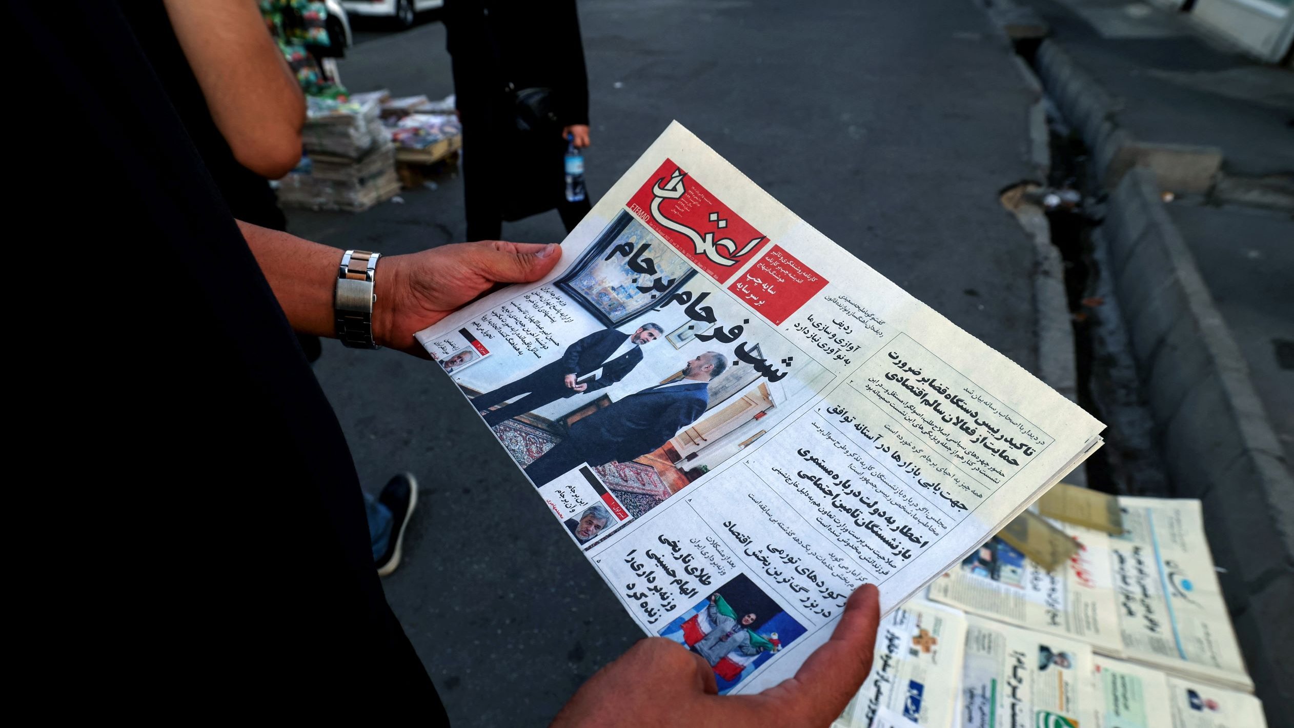 في الصورة، يُظهر شخص يحمل صحيفة باللغة العربية. تبدو الصحيفة جديدة ومطوية، ويظهر عنوان بارز على الصفحه الأولى، كما تُلاحظ مجموعة من الصحف في الخلفية. يبدو أن المشهد يدور في مكان عام، ربما في الشارع، حيث توجد بعض السيارات والأشخاص في محيط الصورة.