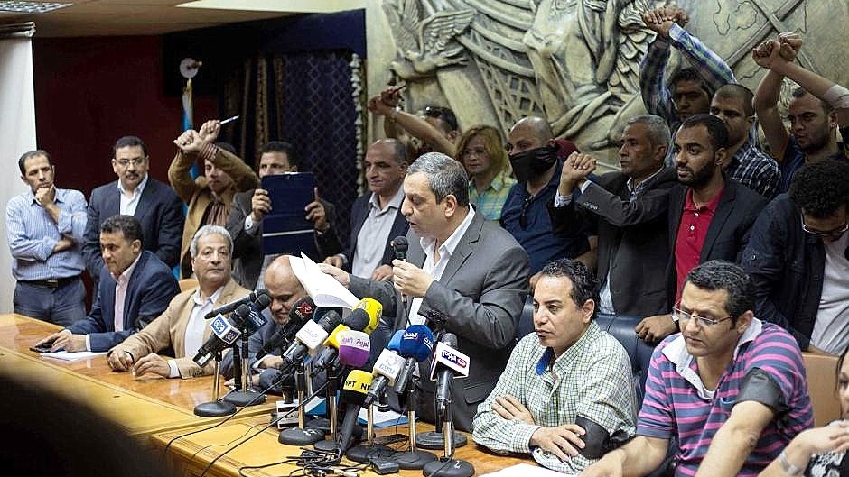 تظهر الصورة اجتماعًا تضامنياً أو مؤتمرًا صحفيًا حيث يجلس عدد من الأشخاص حول طاولة كبيرة، بينما يقف متحدث أمامهم يتحدث إلى الحضور. في الخلفية، يرفع آخرون أيديهم في إشارة إلى الاحتجاج أو التأييد. يظهر البعض في الصورة تفاعلهم مع المتحدث، مما يشير إلى أجواء حماسية. الخلفية تحتوي على منحوتات أو أنماط تزيينية، مما يضيف لمسة رسمية لمكان الاجتماع.