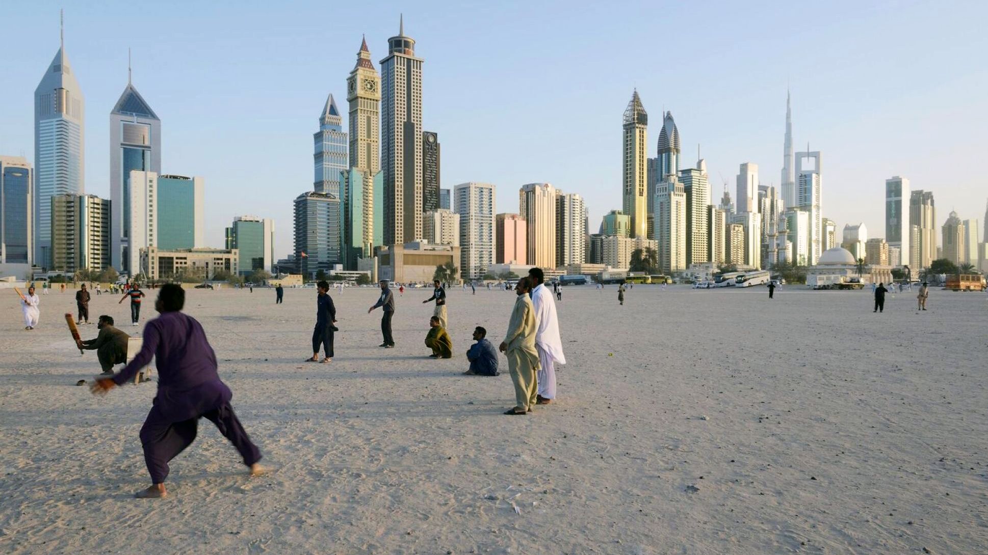 الصورة تُظهر مشهدًا حضريًا لمدينة حديثة، حيث تبرز ناطحات السحاب والمباني الشاهقة. في المقدمة، يوجد مجموعة من الأشخاص يمارسون أنشطة مختلفة على رمال منطقة مفتوحة، ما يوحي بأجواء حيوية. الخلفية تُظهر المدينة مع سماء زرقاء تتخللها أشعة الشمس، مما يضفي لمسة جميلة على المشهد.