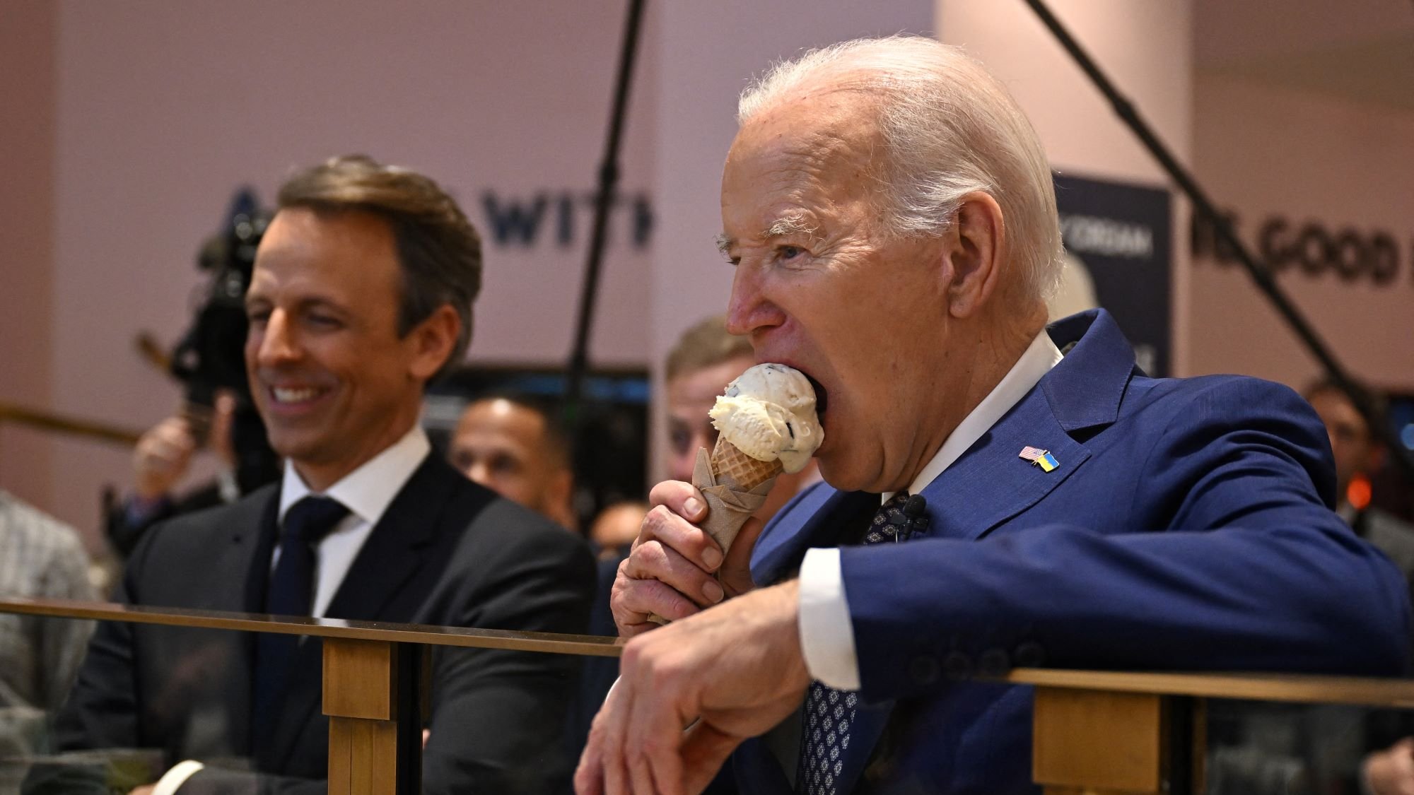 الصورة تُظهر رجلًا في بدلة رسمية وهو يأخذ قضمة من مثلجات، بينما يقف بجانبه رجل آخر يبتسم. يبدو أن المكان عبارة عن حدث أو اجتماع، حيث يوجد العديد من الأشخاص في الخلفية. الرجل الذي يأكل المثلجات هو شخص بارز يبدو سعيدًا وهو يستمتع باللحظة.