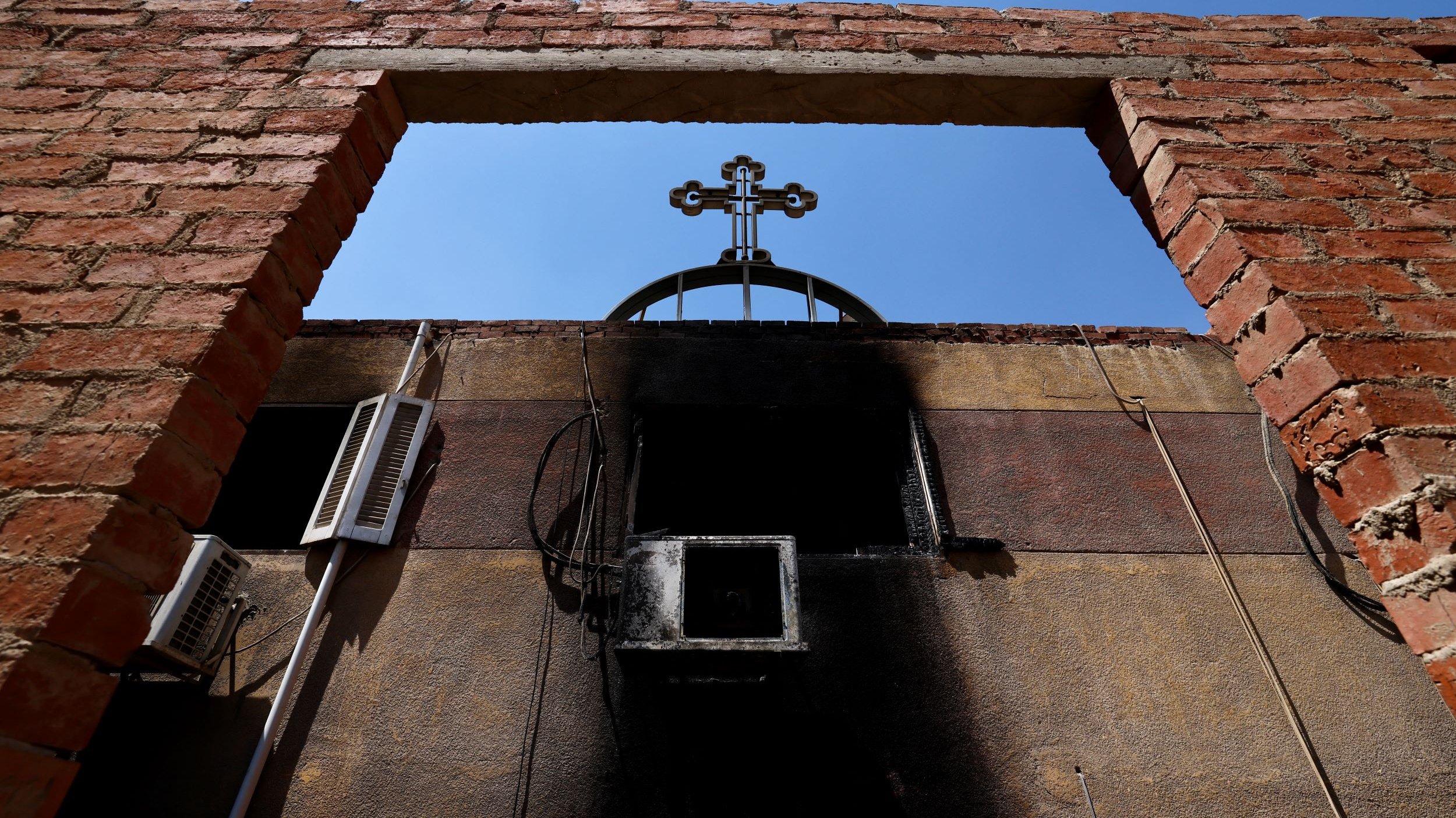 الصورة تظهر إطارًا من الطوب الأحمر مع فتحة في الجزء العلوي، ومن خلال هذه الفتحة يمكن رؤية صليب مرفوع في السماء. الجدران المحيطة تبدو متضررة، حيث يظهر عليها أثر السواد، مما يشير إلى وجود حريق أو تلف. السماء في الخلفية زرقاء وصافية، مما يعكس تناقضًا مع الأجواء المحيطة.