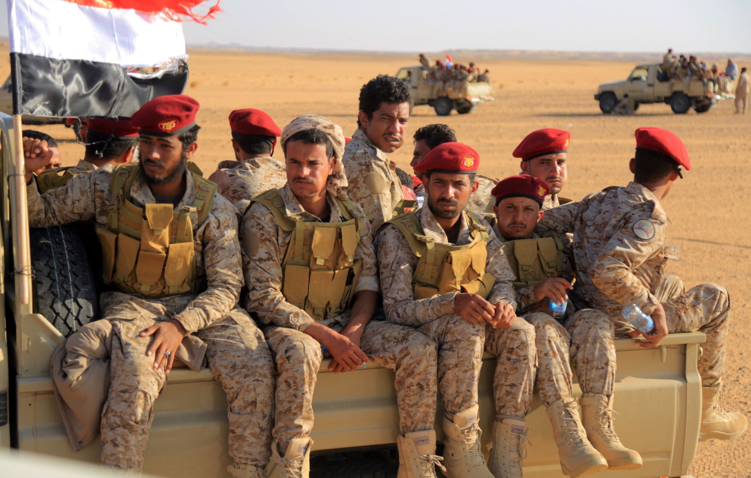 تظهر في الصورة مجموعة من الجنود المرتدين زيًا عسكريًا، يجلسون على مؤخرة مركبة عسكرية في منطقة صحراوية. يرتدي بعض الجنود قبعات حمراء، بينما يظهر العلم اليمني في الخلف. يبدو الجنود في وضعية مريحة، مع تواجد مركبات أخرى في الخلفية. تعكس الصورة أجواء عسكرية في بيئة قاحلة.