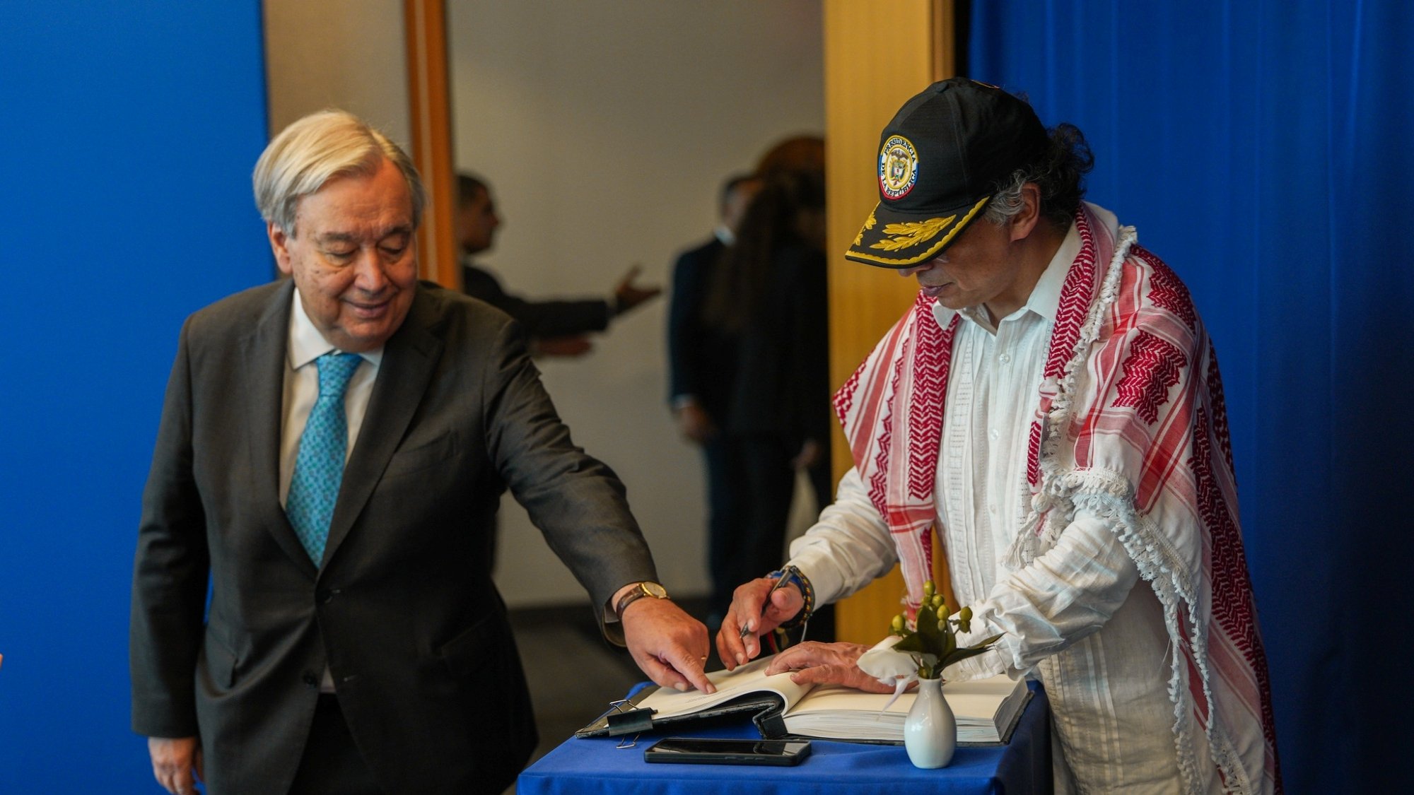 Two men are at a table; one is signing a book while the other observes. A blue backdrop is behind them.