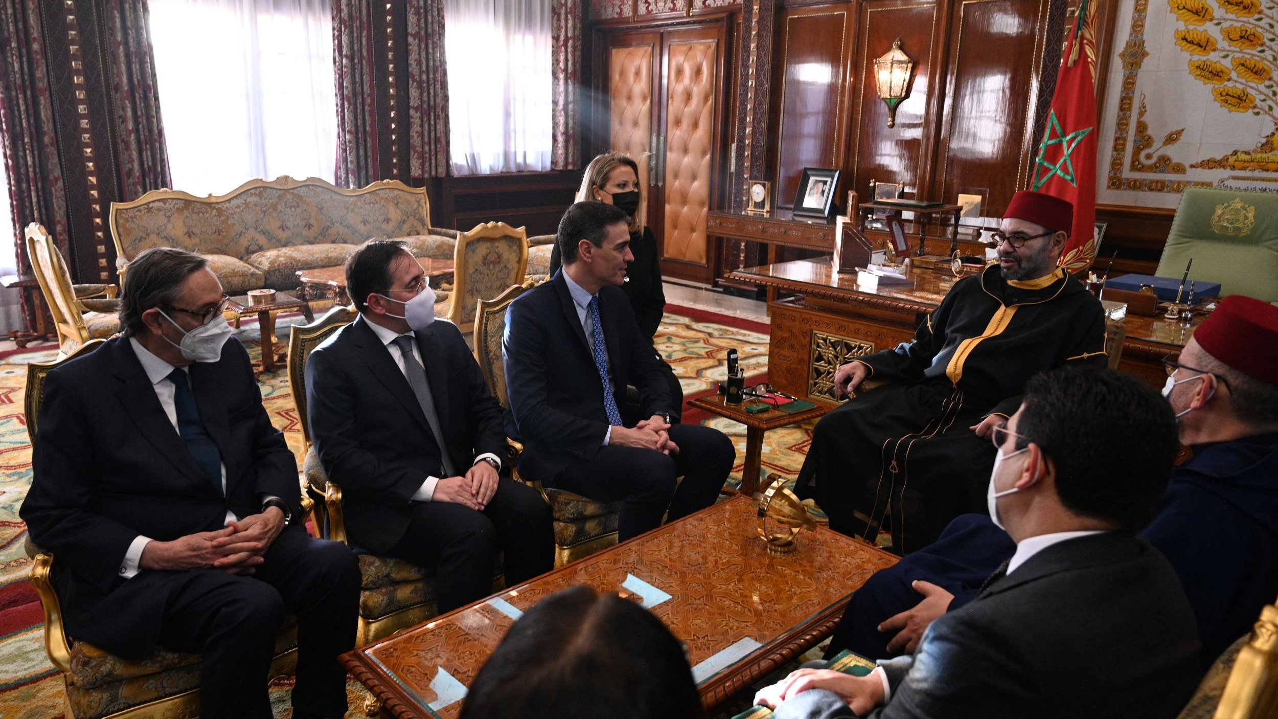 تظهر الصورة مجموعة من الأشخاص يجلسون في غرفة ذات تصميم تقليدي مغربي جميل. يجلس في المنتصف شخص يرتدي لباسًا رسميًا، بينما يجلس الآخرون حوله في مقاعد مريحة. يبدو المكان مزخرفًا بألوان دافئة ونقوش معمارية، مع وجود نافذة كبيرة تسمح بدخول الضوء الطبيعي. شخصياتهم تعكس اهتمامًا بالحديث، ويظهرون بملابس رسمية، مما يشير إلى حدوث اجتماع أو مناقشة مهمة.