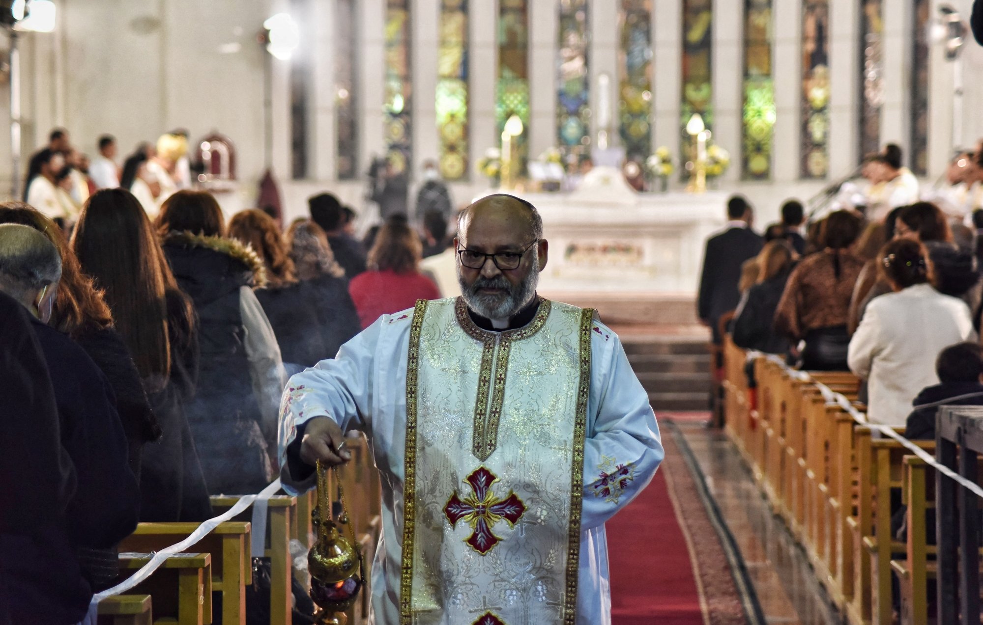 A priest in white robes walks down the aisle of a church during a service.