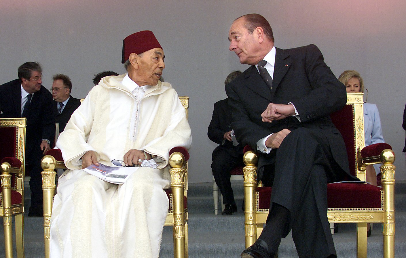 الصورة تظهر شخصين يجلسان على كراسي فاخرة. يبدو أن الحديث يجري بينهما بشكل ودي. الشخص الأول يرتدي زيًا تقليديًا مغربيًا مع طربوش، بينما الشخص الثاني يرتدي بدلة رسمية داكنة. في الخلفية توجد مجموعة من الأشخاص الجالسين، ويبدو أنهم في مناسبة رسمية أو احتفال. الضوء في الصورة ناعم، مما يضيف جوًا من الألفة.