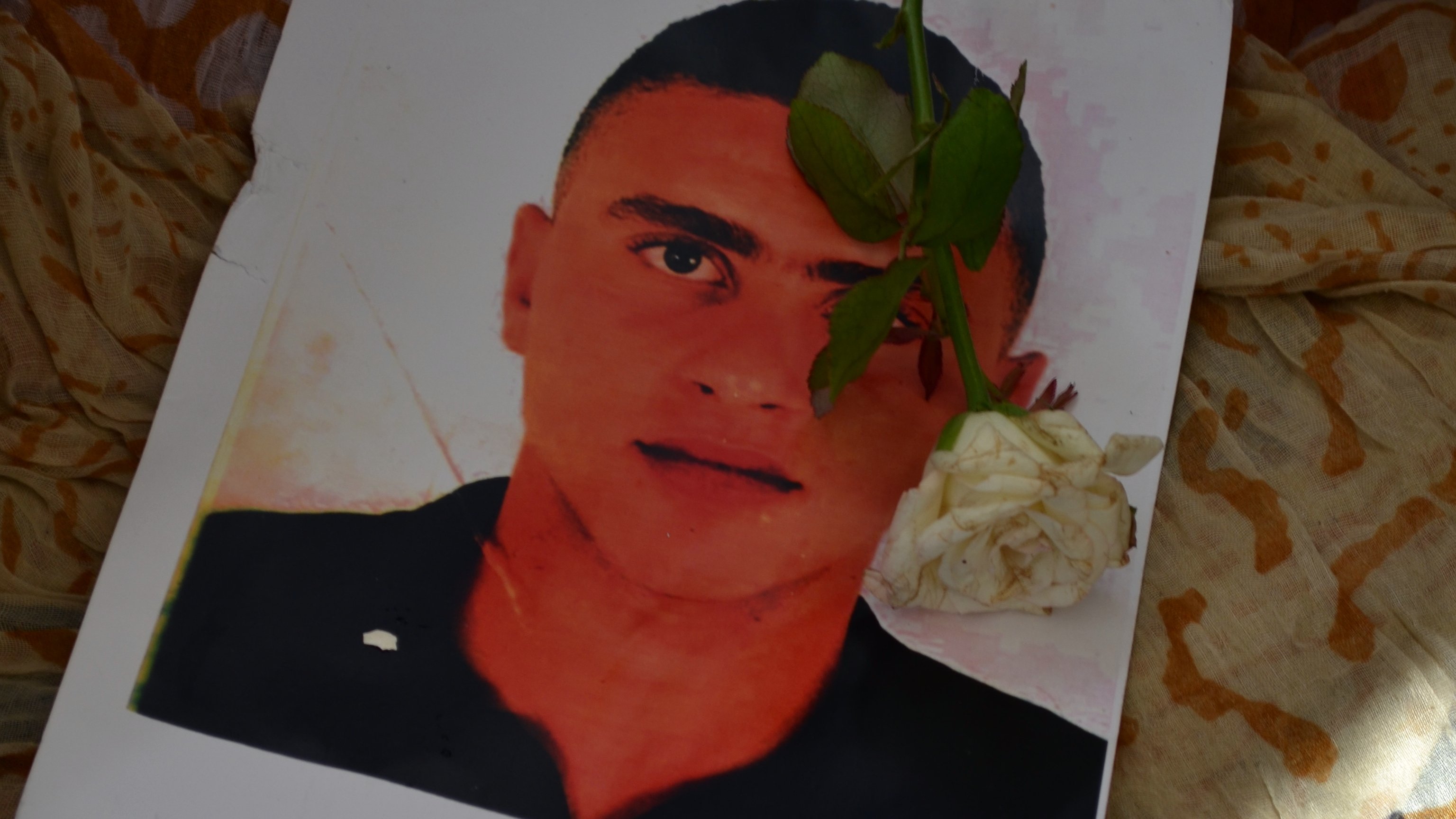 تظهر في الصورة ورقة مطبوعة تحتوي على صورة شاب، حيث يوضع عليها وردة بيضاء. الخلفية تبدو ملونة ومعقدة، مما يضيف جوًا من الحزن والحنين. الورقة قد تشير إلى ذكرى لهذا الشخص، والزهرة تعزز من مشاعر التأمل والاحترام.