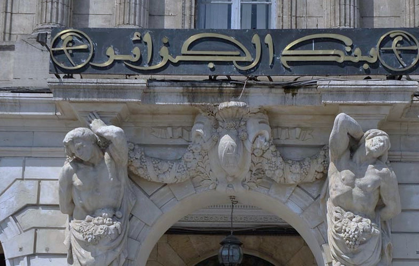 تظهر الصورة واجهة مبنى، تحمل لافتة واضحة مكتوب عليها "بنك الجزائر". يتميز المدخل بتفاصيل معمارية مميزة، حيث يحيط به تماثيل لأشخاص يحملون زينة أو ثمار، مما يضيف لمسة فنية وجمالية للواجهة. يُظهر التصميم العناية بالتفاصيل والزخارف، مما يعكس الطراز المعماري الفريد للمكان.