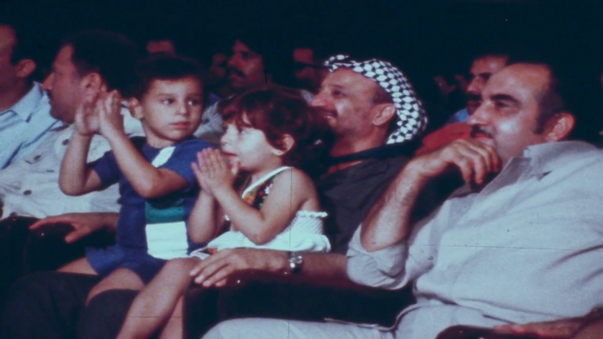 في الصورة مجموعة من الأشخاص يجلسون في قاعة، ومن بينهم طفلان يجلسان أمام رجل يرتدي كوفية. يبدو أن الجميع يستمع أو يشاهد شيئًا ما في الفعالية. الأطفال يظهرون تفاعلهم بالتصفيق أو الاستمتاع بالعرض. الرجال في الصورة يبدون مهتمين ومسترخين. الجو يبدو احتفاليًا أو حيويًا.