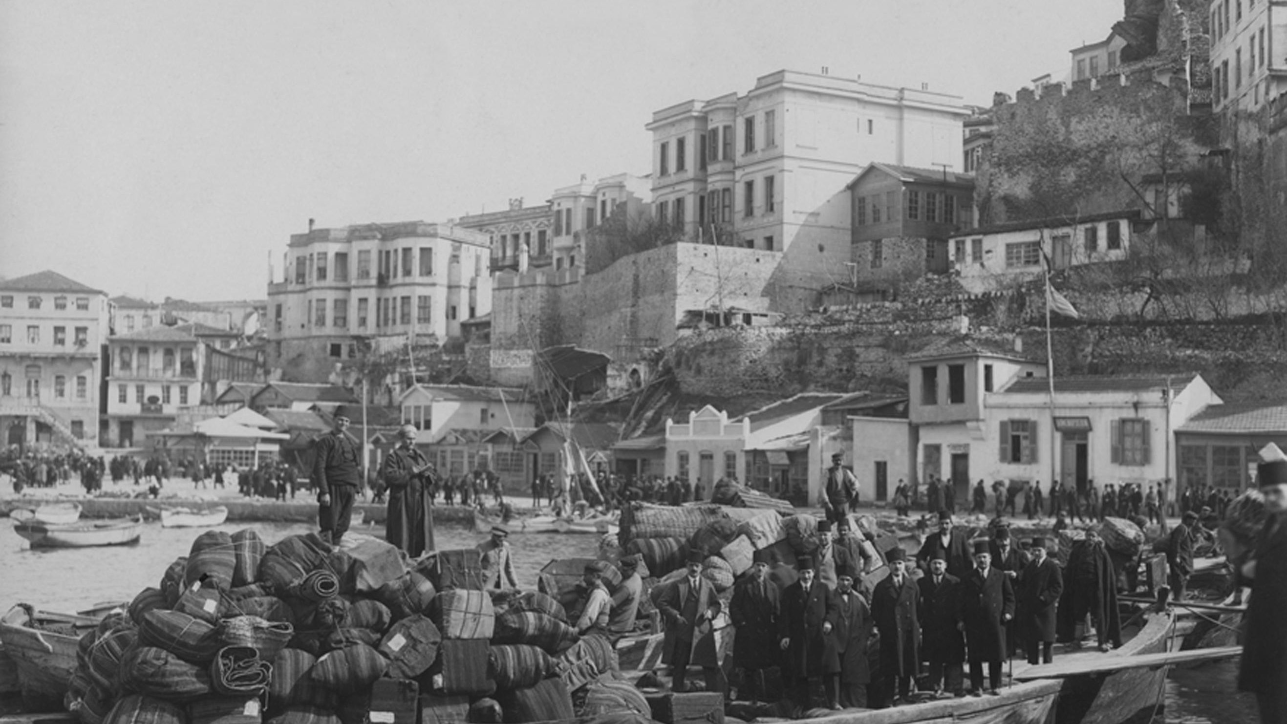 تظهر الصورة مشهداً تاريخياً من منطقة ساحلية، حيث توجد مجموعة من الأشخاص يقفون على قارب محمّل بأكياس وصناديق، مما يدل على حركة تجارية أو نقل للبضائع. في الخلفية، يمكن رؤية مبانٍ متعددة الطوابق وبعض الأسطح، بالإضافة إلى قلعة أو حصن يبدو أنه في حالة جيدة. هناك مجموعة من الناس تسير على الرصيف، مما يدل على النشاط والحركة في المنطقة. الأجواء العامة تعكس فترة زمنية سابقة، ربما أوائل القرن العشرين، مع تنوع في الملابس التي تشير إلى زمن بعيد.