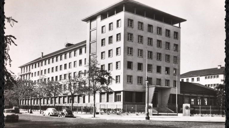 الصورة تُظهر مبنىً حديث التصميم، يتكون من عدة طوابق وله واجهة متنوعة الألوان. يتواجد حول المبنى مجموعة من الأشجار، ويبدو أن هناك شارعًا أمام المبنى حيث توجد بعض السيارات القديمة. يعكس تصميم المبنى طابعًا معماريًا بسيطًا وعصريًا، مما يوحي بأنه تم بنائه في فترة زمنية معينة في القرن العشرين. السماء تبدو واضحة مما يعطي انطباعًا بأن الصورة قد تم التقاطها في يوم مشمس.