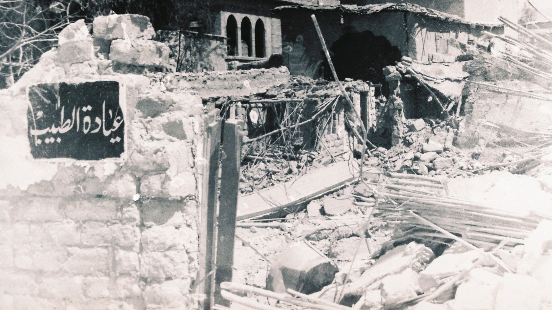 الصورة تُظهر مشهداً لمدمرات بسبب أحداث سابقة، حيث يظهر حطام المباني والخرائب. في مقدمة الصورة، توجد لافتة تحمل اسم مكان، ولكنها مهملة وسط الأنقاض. الخلفية تحتوي على نوافذ وأجزاء من مباني قديمة، مما يعكس تأثير الزمن أو كارثة معينة. الأجواء توحي بالحزن والألم الناتج عن الدمار.