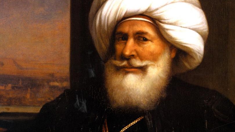 تظهر الصورة رجلاً مسنًّا يرتدي عمامة كبيرة ومجموعة من الملابس التقليدية. يتميز بلحيته البيضاء الكثيفة وابتسامة ودودة. يحمل سيفًا في يده، مما يوحي بمكانته العالية أو دوره القيادي. الخلفية تحتوي على مشهد يبدو كأنه منظر طبيعي لمدينة أو تجمع حضري. تعكس تفاصيل ملابسه وزينه التراث الثقافي لمنطقته.