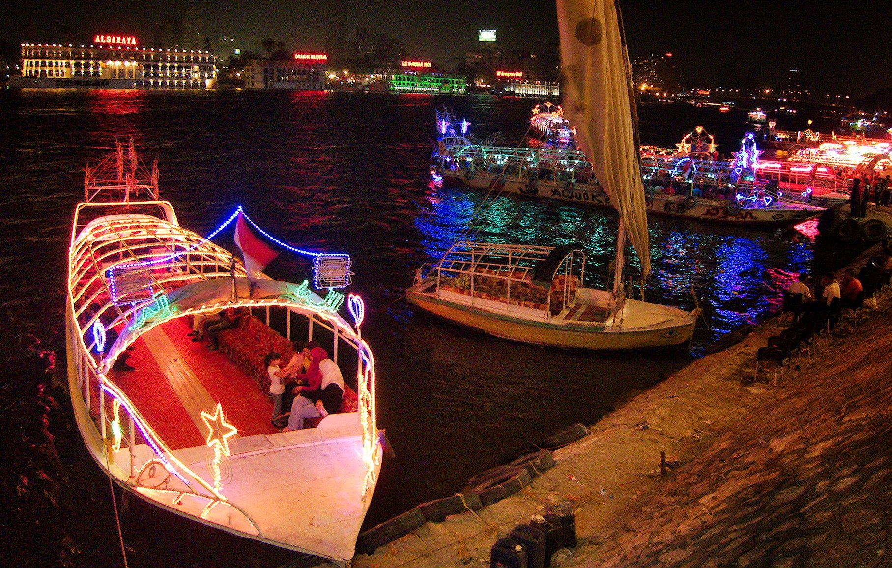 تظهر الصورة مشهدًا ليليًا لنهر، حيث تضيء القوارب بأضواء ملونة وزاهية. يمكن رؤية قوارب أخرى في الخلفية، وبعض الناس يجلسون على ضفاف النهر. تتميز الأجواء بألوان مشرقة تعكس أجواء من الاحتفال والمرح.